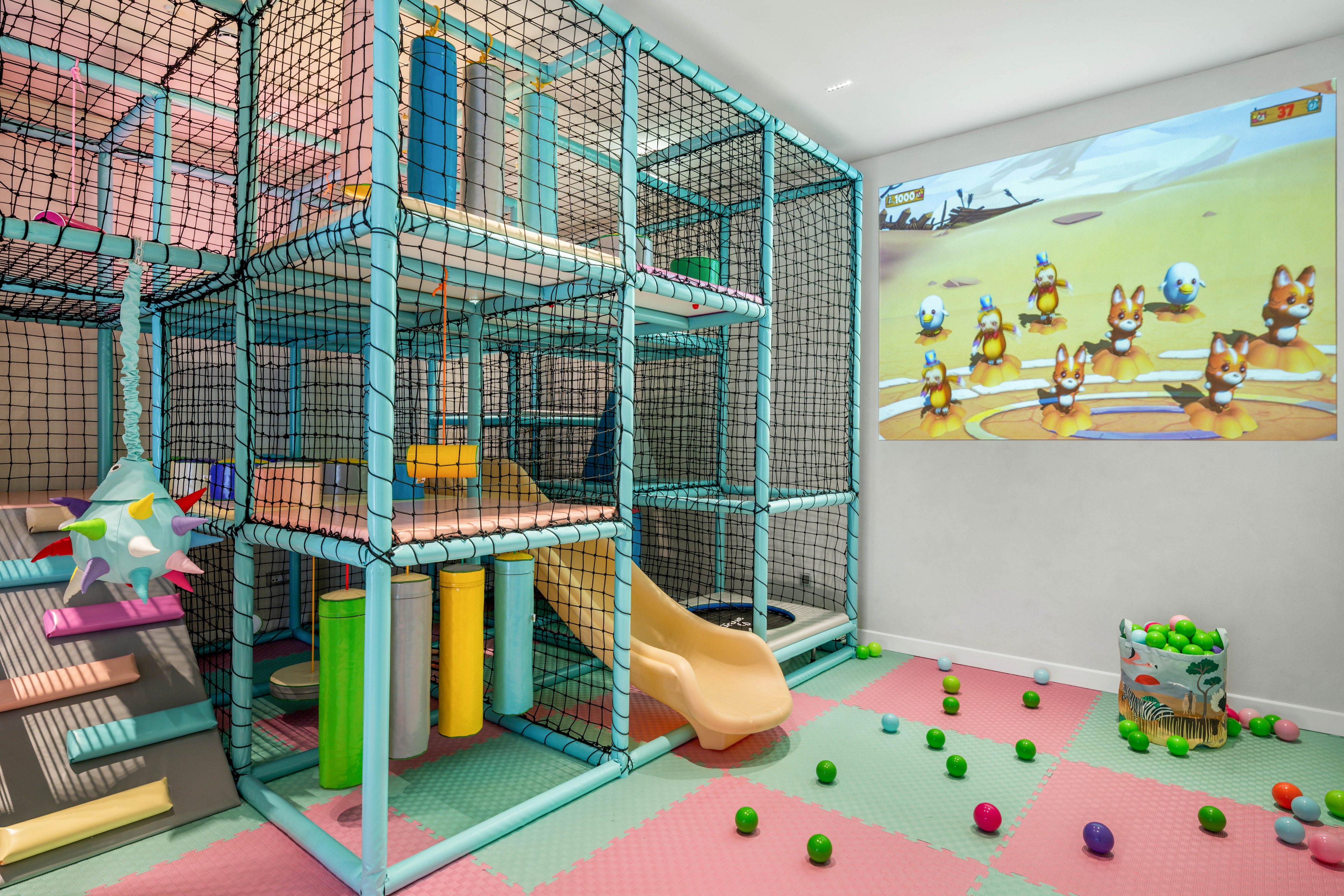Colorful indoor playroom features a multi-level soft play structure with padded platforms and a small slide. The floor is covered in pastel foam tiles with scattered plastic balls and soft play elements. A large wall projection shows an animated game with cartoon animals and soccer balls. The space appears bright, safe, and designed for young children’s active play.