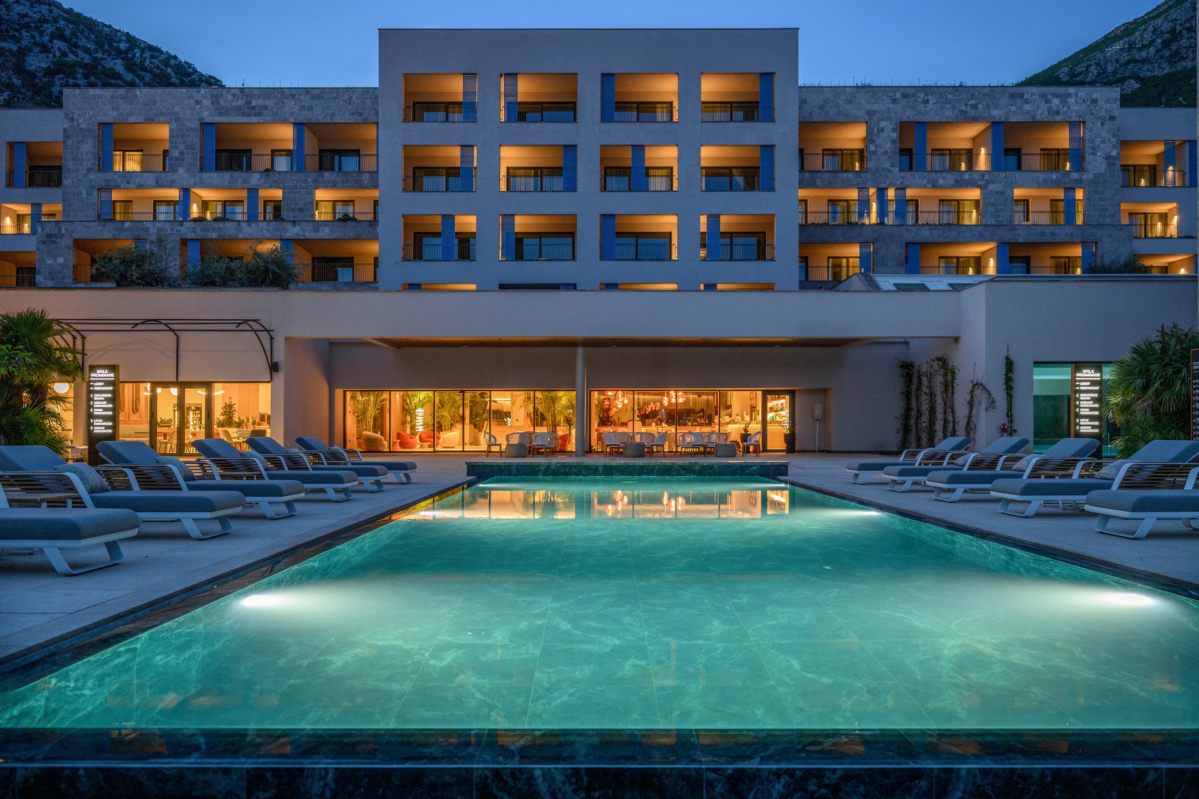 A contemporary multi-story hotel stands behind a large illuminated swimming pool at dusk. Lounge chairs line both sides of the pool, facing the building’s balconies and terraces. Warm interior lighting from the lobby and bar contrasts with the cool blue water and twilight sky. The surrounding mountains frame the resort, emphasizing a tranquil vacation setting.