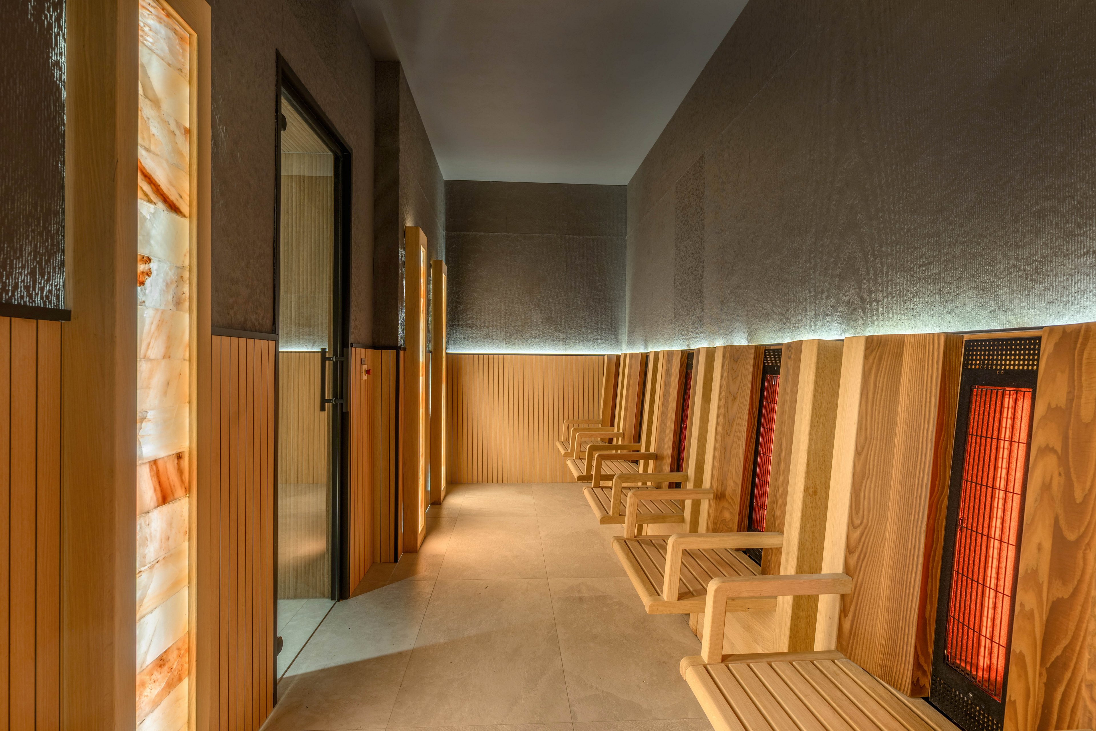 A contemporary sauna room features a row of wooden reclining seats along a narrow corridor. Infrared heating panels and illuminated salt or stone columns line the walls, creating a warm, inviting glow. The space is finished with light wood paneling, textured upper walls, and soft indirect lighting. A glass door at the end of the corridor suggests access to additional spa facilities.