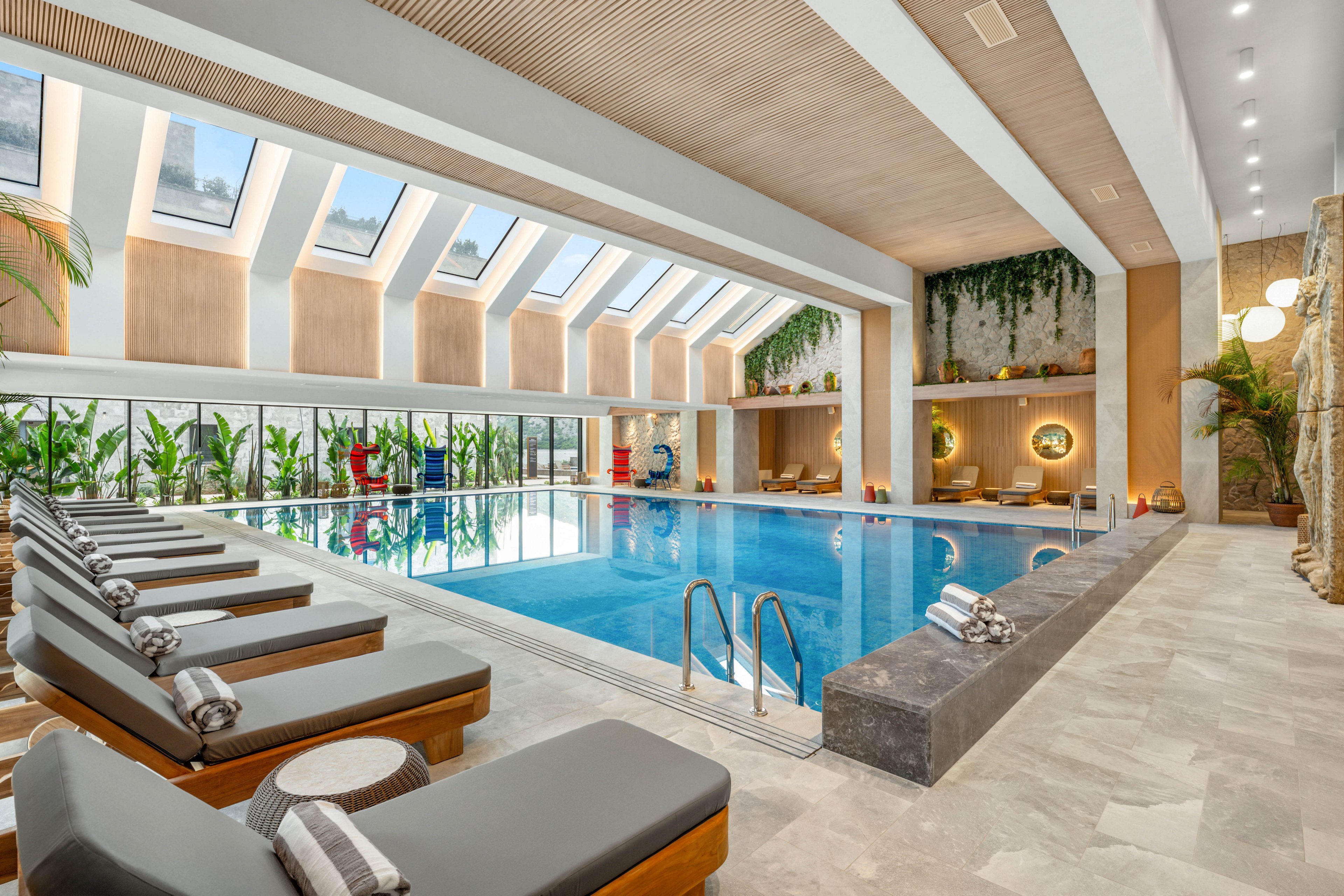 Bright indoor swimming pool area featuring multiple cushioned loungers with rolled towels. Large skylights and floor-to-ceiling windows fill the space with natural light and views of greenery. Warm wood accents, stone flooring, and hanging plants create a modern spa-like atmosphere. Soft ambient lighting and decorative wall niches enhance the relaxing, upscale setting.