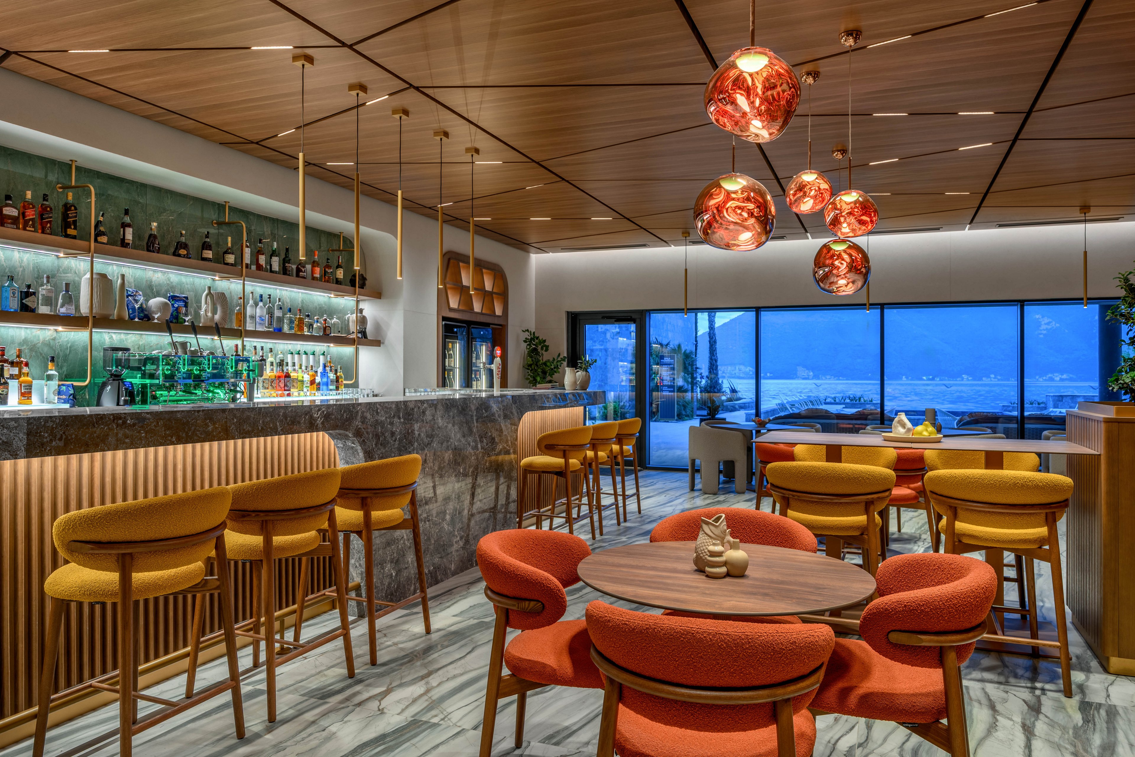 Contemporary bar interior featuring a marble counter and illuminated liquor shelves. Warm orange chairs and stools surround wooden tables under copper pendant lights. Large floor-to-ceiling windows reveal an evening ocean view with outdoor seating. The space combines sleek architectural lines with cozy, upscale lounge decor.