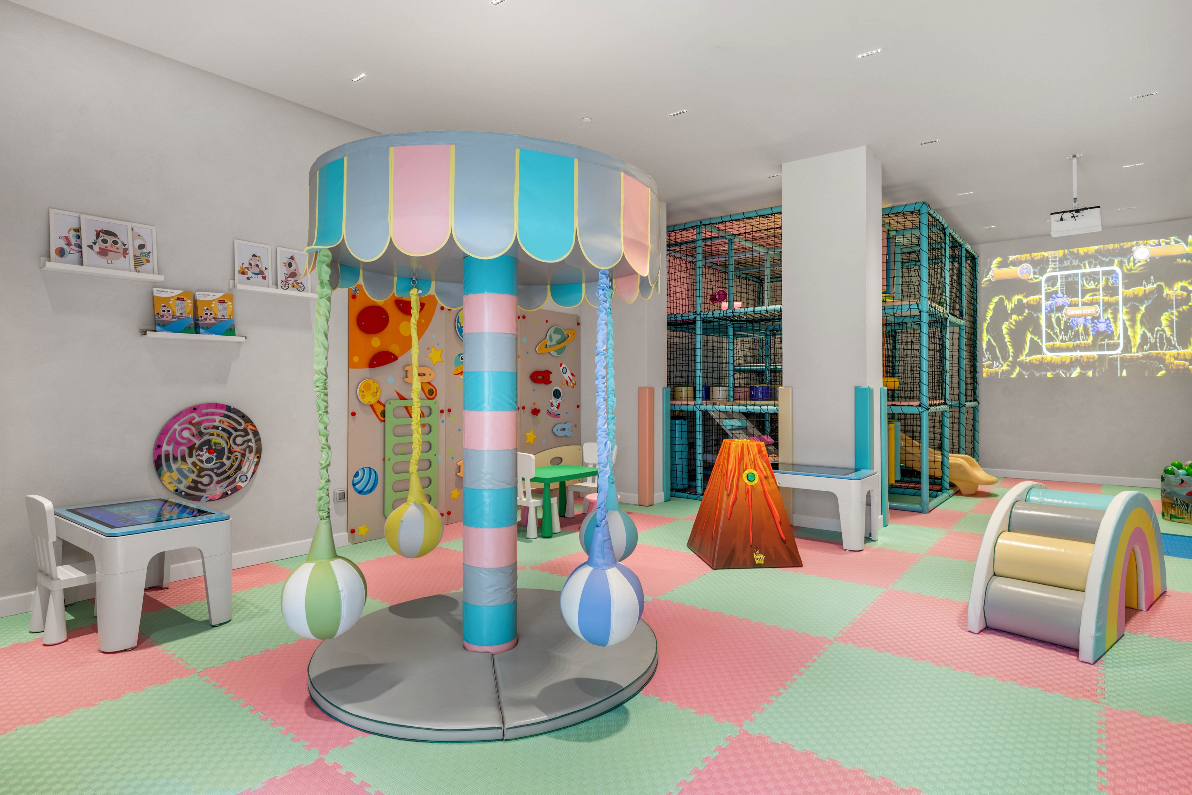 Brightly colored indoor playroom features a central pastel carousel-style play structure with hanging soft balls. The space includes foam floor tiles in pink, green, and light blue, along with child-sized tables, chairs, and interactive play equipment. A climbing area with safety netting stands in the background, while a rainbow-shaped tunnel and cone-shaped slide add playful variety. Wall art and illuminated line drawings contribute to the cheerful, modern atmosphere designed for young children.