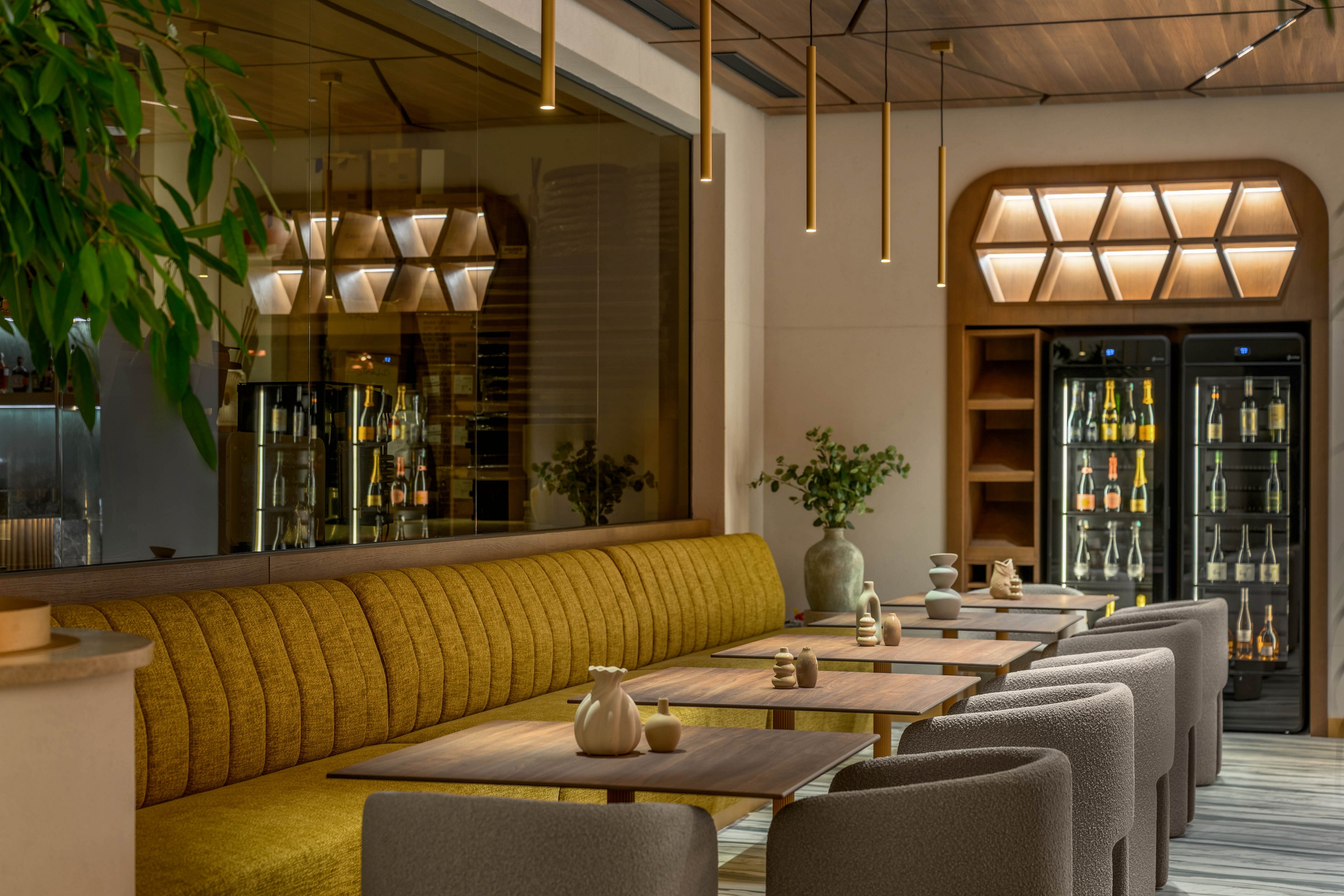 Contemporary restaurant interior featuring a long mustard-yellow upholstered bench and light gray chairs around wooden tables. In the background, a glass-front refrigerator displays bottles of beverages, adding depth and interest to the space. Warm pendant lighting and geometric wall lights create a soft, inviting ambiance. Decorative plants and minimalist tableware enhance the modern, stylish atmosphere.