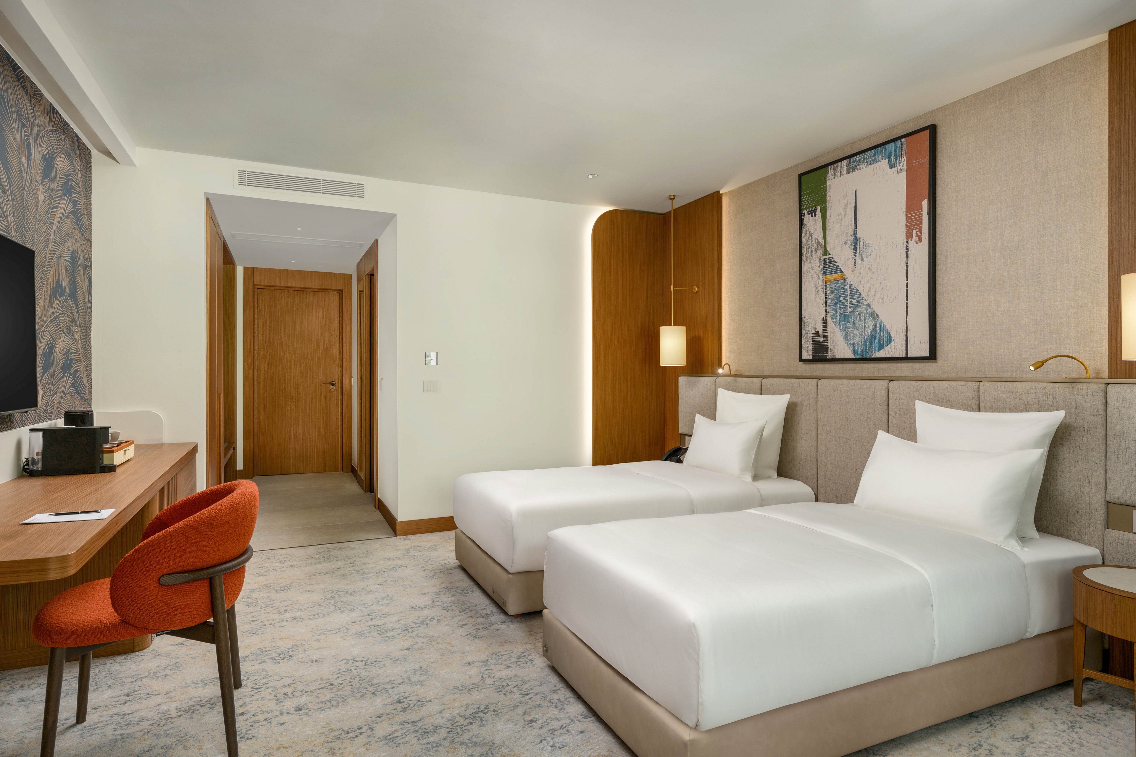 Contemporary hotel guest room featuring two neatly made twin beds with white linens. The space includes a wooden desk with an orange upholstered chair and a doorway leading to the hall. Soft neutral tones, warm wood accents, and abstract wall art create a calm, inviting atmosphere. Subtle lighting and a clean, uncluttered layout emphasize comfort and modern design.