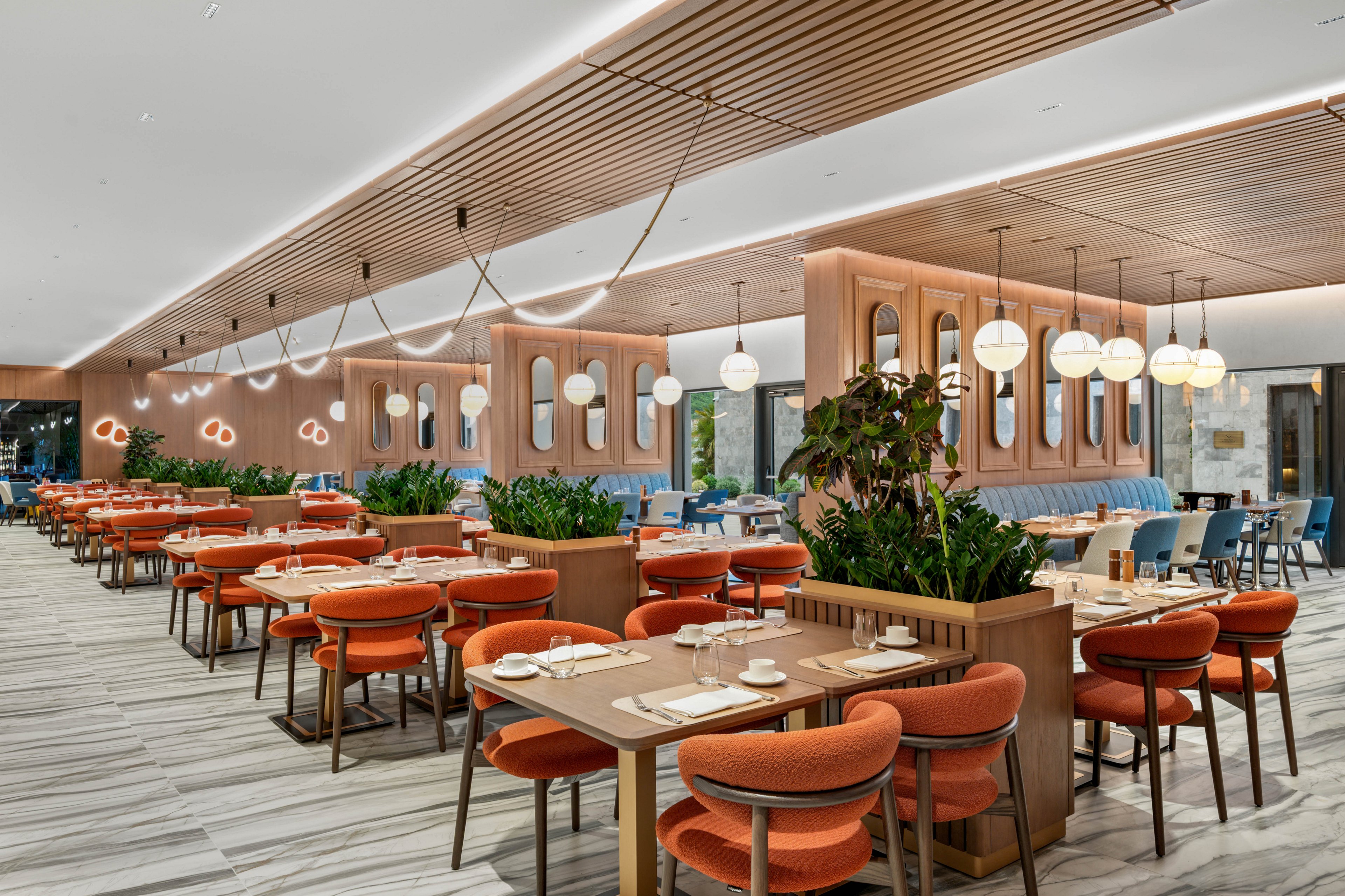 Contemporary restaurant interior features rows of wooden tables with bright orange upholstered chairs. The spacious dining area is accented by large planters with green foliage and a light wood slatted ceiling. Round white pendant lights and wall mirrors create a stylish, upscale atmosphere. The space appears ready for service with neatly arranged place settings and a clean, open layout.