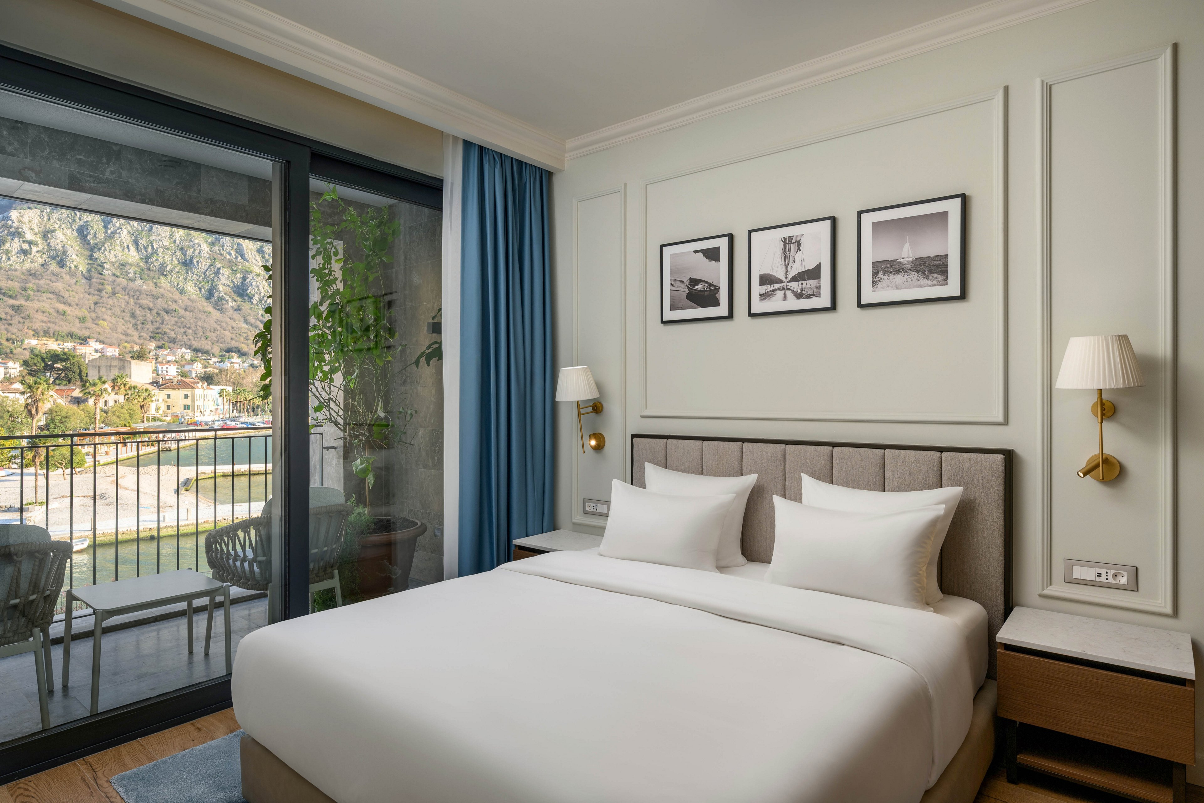 Contemporary hotel room featuring a neatly made double bed with white linens and upholstered headboard. A large glass sliding door opens to a balcony with outdoor chairs and a scenic mountain and town view. The interior includes framed black-and-white wall art, bedside tables with wall-mounted lamps, and blue curtains framing the window. Soft neutral tones and natural light create a calm, inviting atmosphere suitable for relaxation or vacation stays.