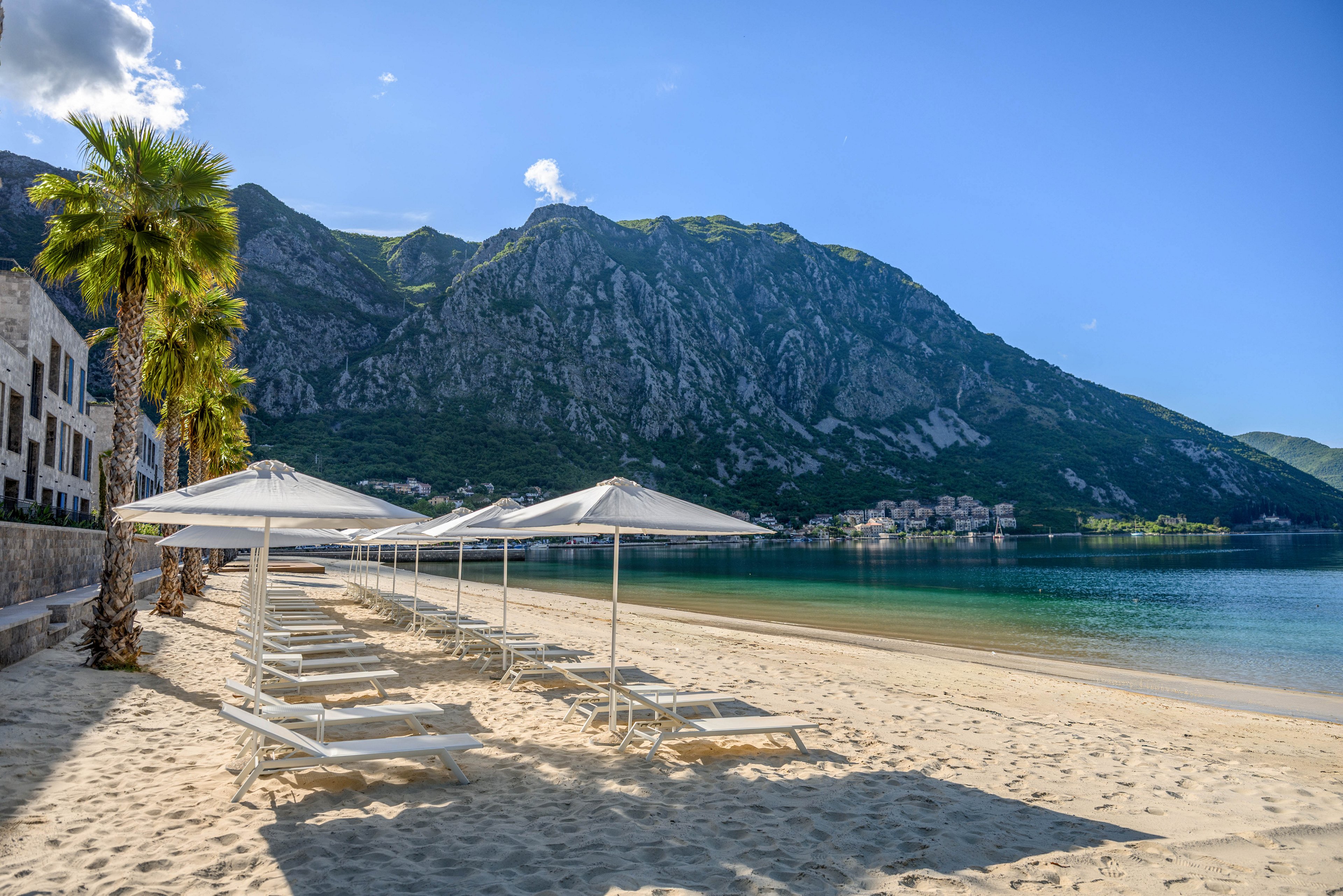 A row of white sun loungers and umbrellas stands on a sandy beach beside calm, clear water. Palm trees line the edge of the beach near white buildings on the left. In the background, steep green mountains rise dramatically under a bright blue sky with scattered clouds. The scene feels peaceful and inviting, suggesting a quiet seaside resort.