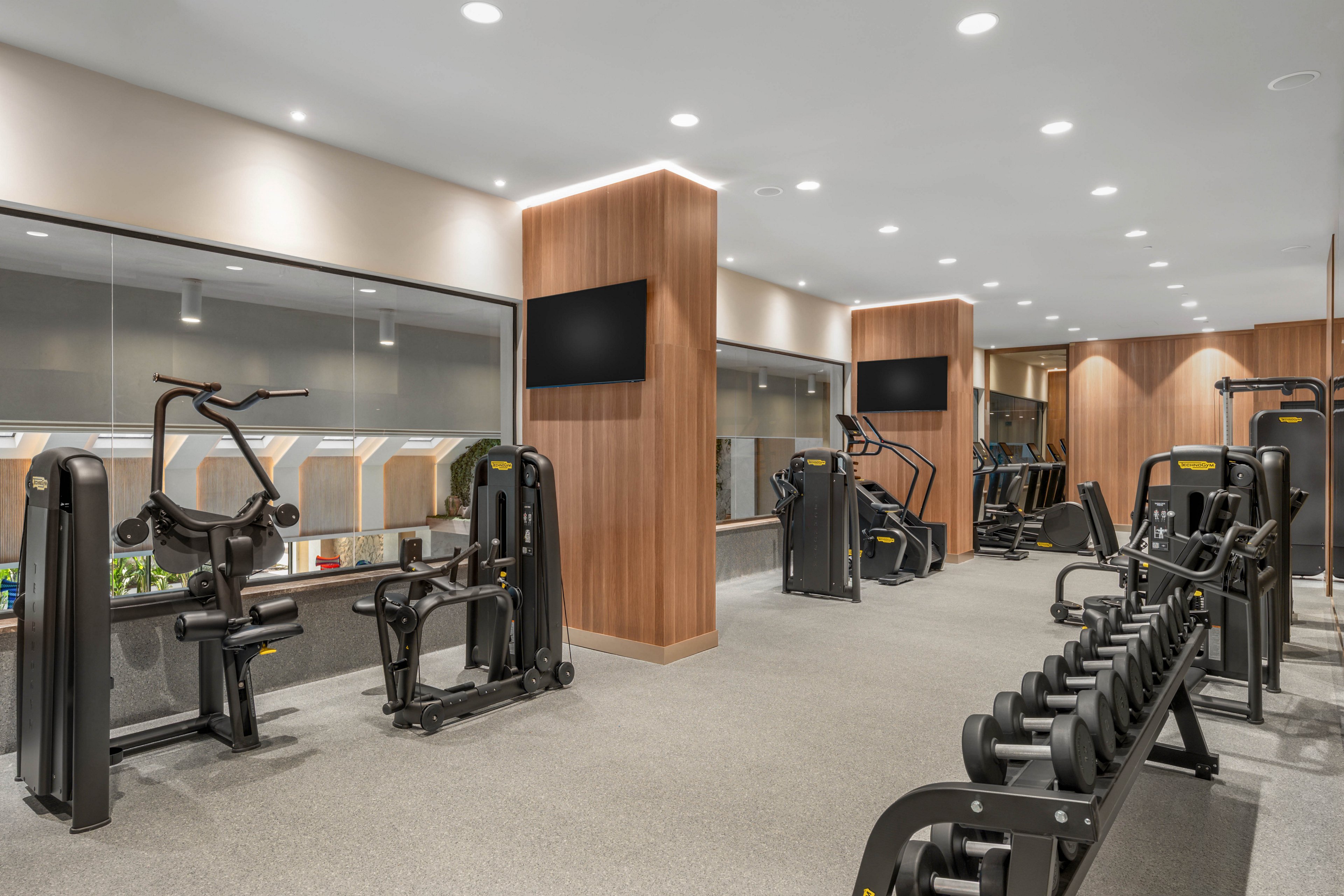 A contemporary fitness center features rows of strength training machines and free weights. Wooden wall panels and recessed ceiling lights create a clean, upscale atmosphere. Large windows look out onto another interior space, while flat-screen monitors are mounted on central columns. The gym appears empty, emphasizing the equipment and spacious layout.