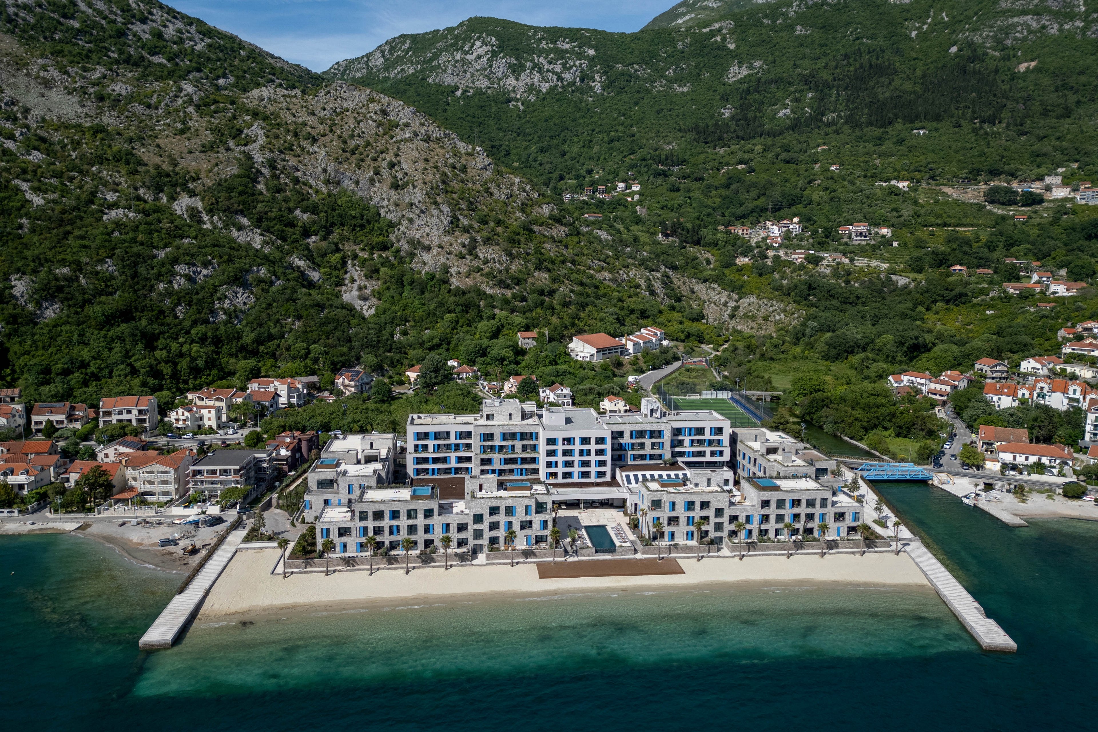 A large contemporary seaside resort stretches along a sandy beach beside clear turquoise water. The complex features white and blue facades, multiple terraces, and pools facing the sea. It is surrounded by smaller coastal houses and backed by steep, forested mountains. The aerial perspective highlights the resort’s scale and its scenic coastal setting.