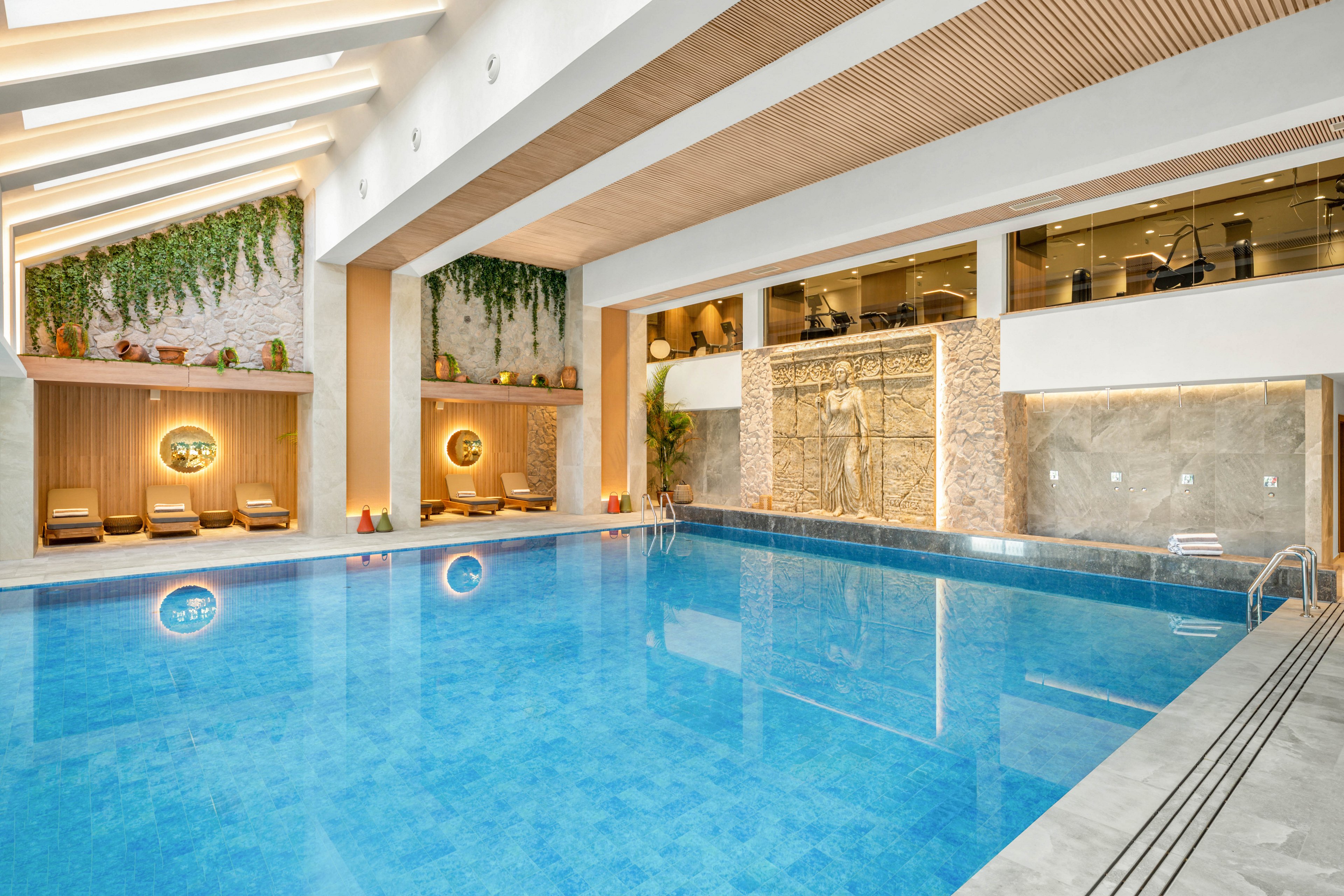 Bright indoor swimming pool in a modern, upscale facility with clear blue water. Comfortable lounge chairs and circular wall lights line a wooden alcove at one end of the pool. Green hanging plants and large stone-textured walls add a natural, spa-like atmosphere. Glass windows reveal an upper-level fitness or relaxation area, enhancing the sense of spaciousness and luxury.