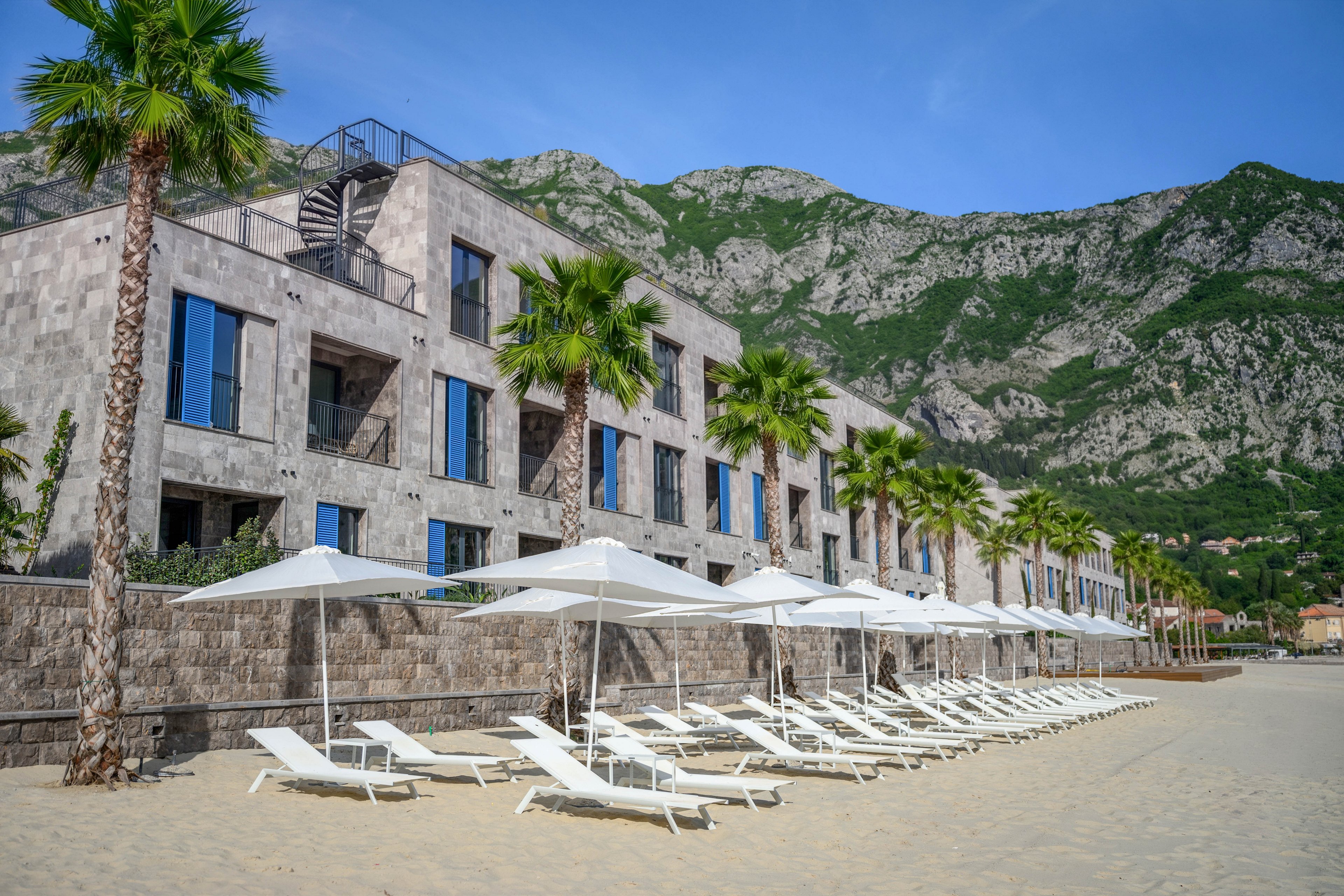 A contemporary stone-clad resort building faces a sandy beach lined with white sun loungers and umbrellas. Tall palm trees stand between the hotel and the shoreline, adding a tropical feel to the scene. Rugged green mountains rise dramatically in the background under a clear blue sky. The overall atmosphere is calm and inviting, suggesting a relaxing seaside vacation destination.