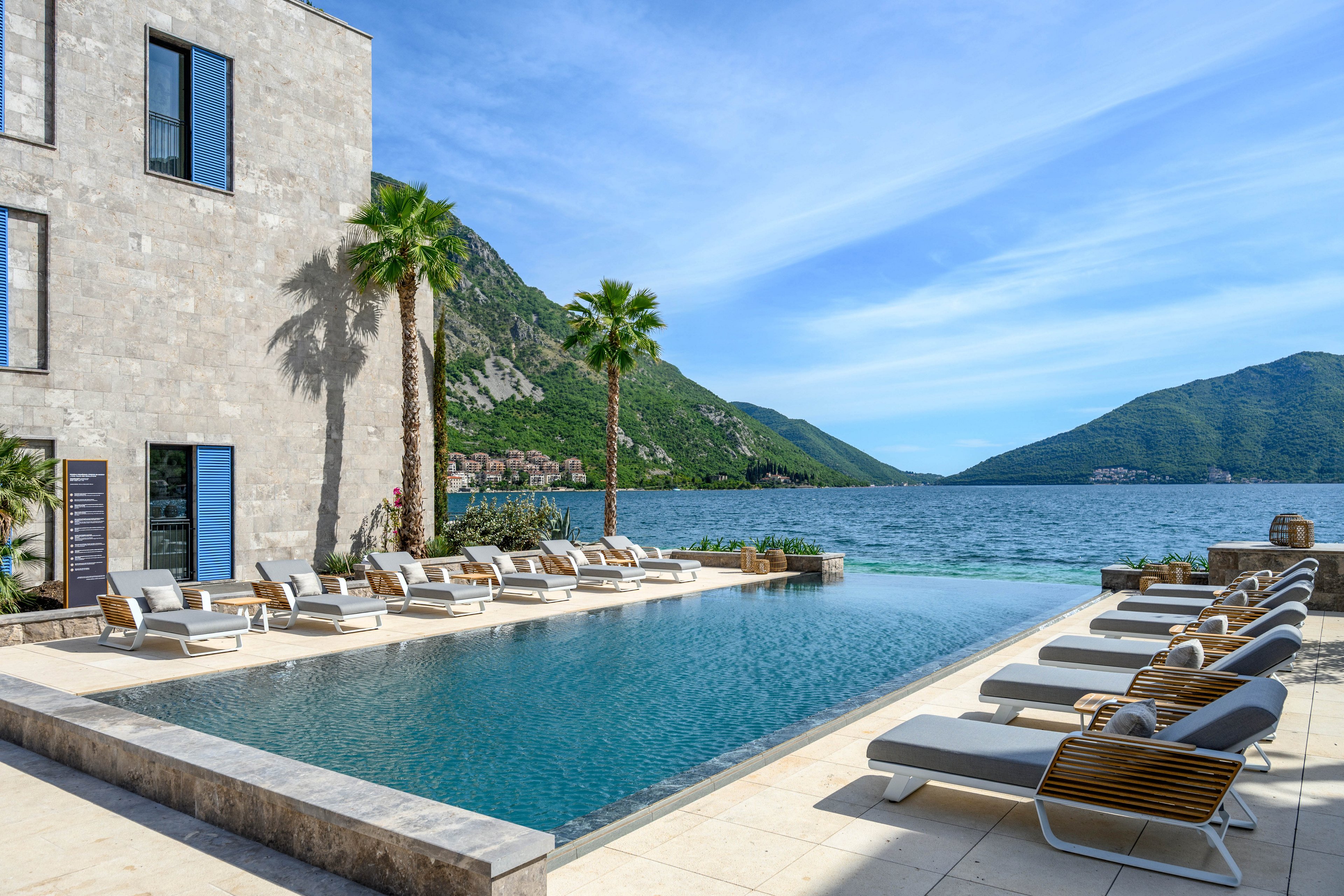 A modern infinity pool stretches toward a calm blue sea, framed by a stone hotel building and tall palm trees. Numerous sun loungers with cushions line the poolside, suggesting a relaxing resort atmosphere. Green mountains rise in the background under a clear, bright sky, enhancing the scenic coastal setting. The overall mood is tranquil, sunny, and inviting for vacation or leisure.
