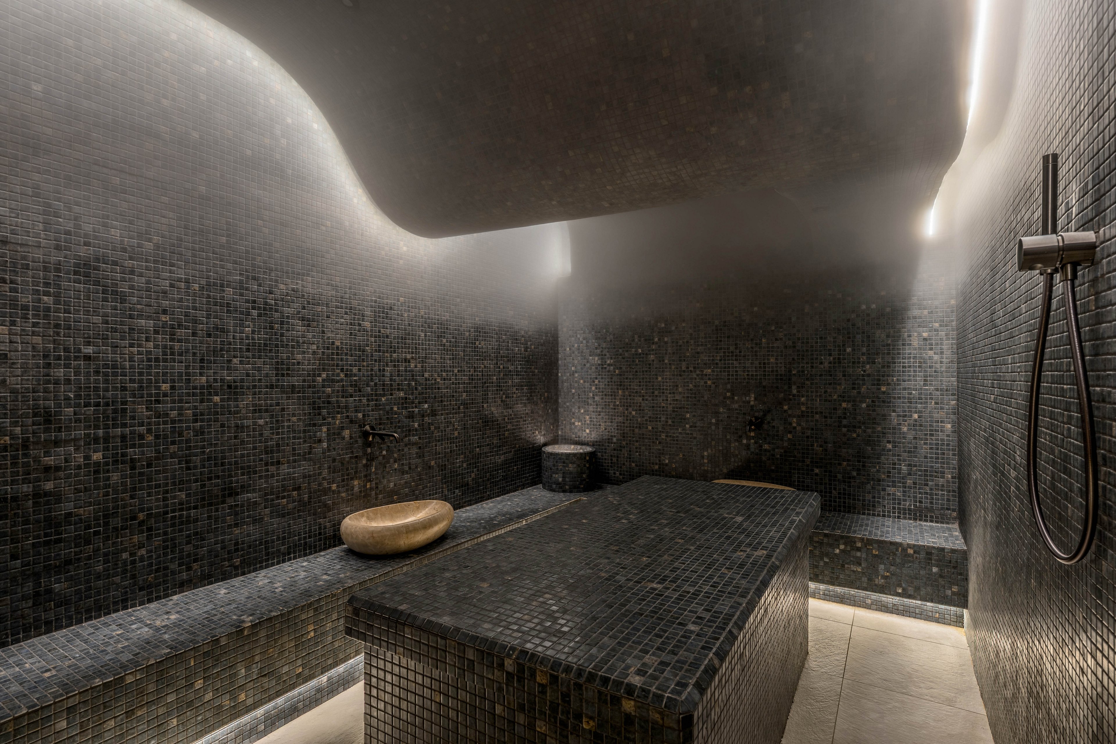 A contemporary steam room features dark mosaic tiles covering the benches, walls, and central massage table. Soft steam rises from the back wall, creating a relaxing spa atmosphere. A smooth stone basin sits on the tiled surface, and a sleek wall-mounted shower fixture is visible on the right. Subtle linear lighting highlights the curves of the ceiling and the clean, minimalist design.