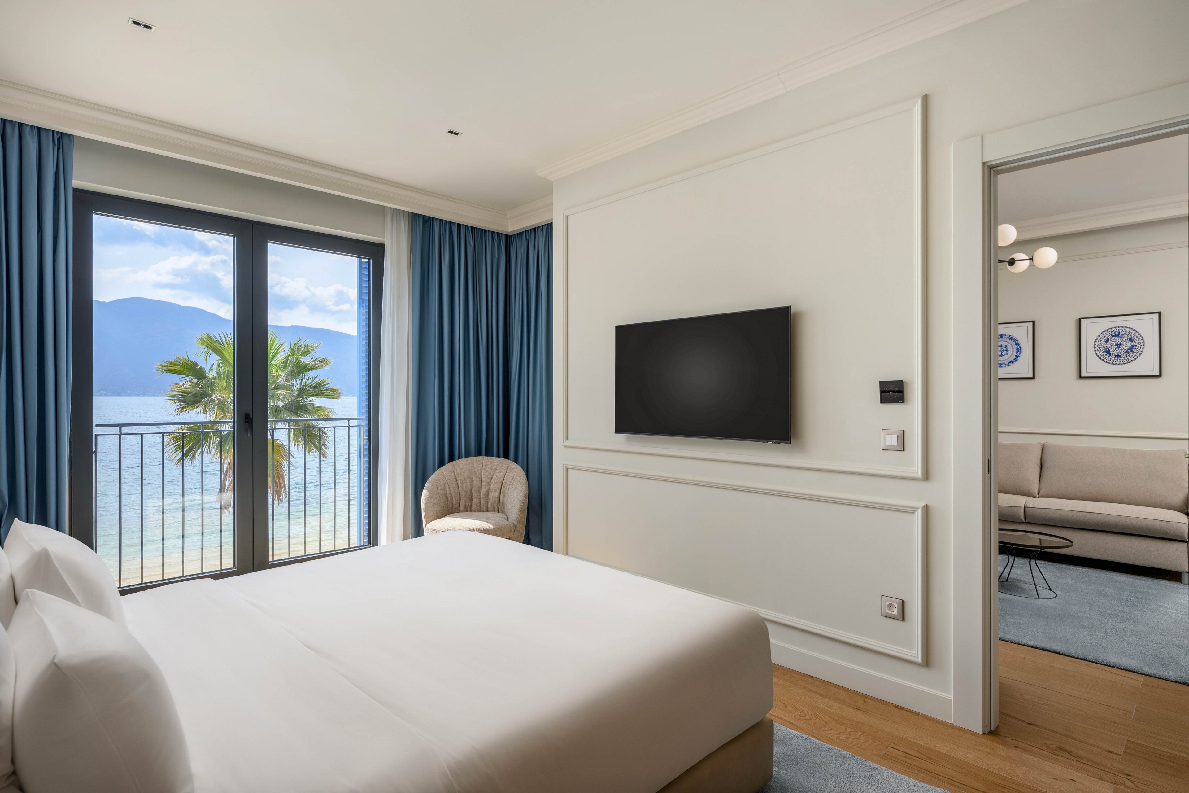 A contemporary hotel bedroom features a neatly made bed facing a wall-mounted flat-screen television. Large glass doors open onto a balcony with a view of the sea, mountains, and a palm tree. Blue curtains and a soft armchair add a calm, coastal feel to the space. An adjoining sitting room with a sofa and framed artwork is visible through an open doorway, enhancing the sense of a spacious, relaxing suite.