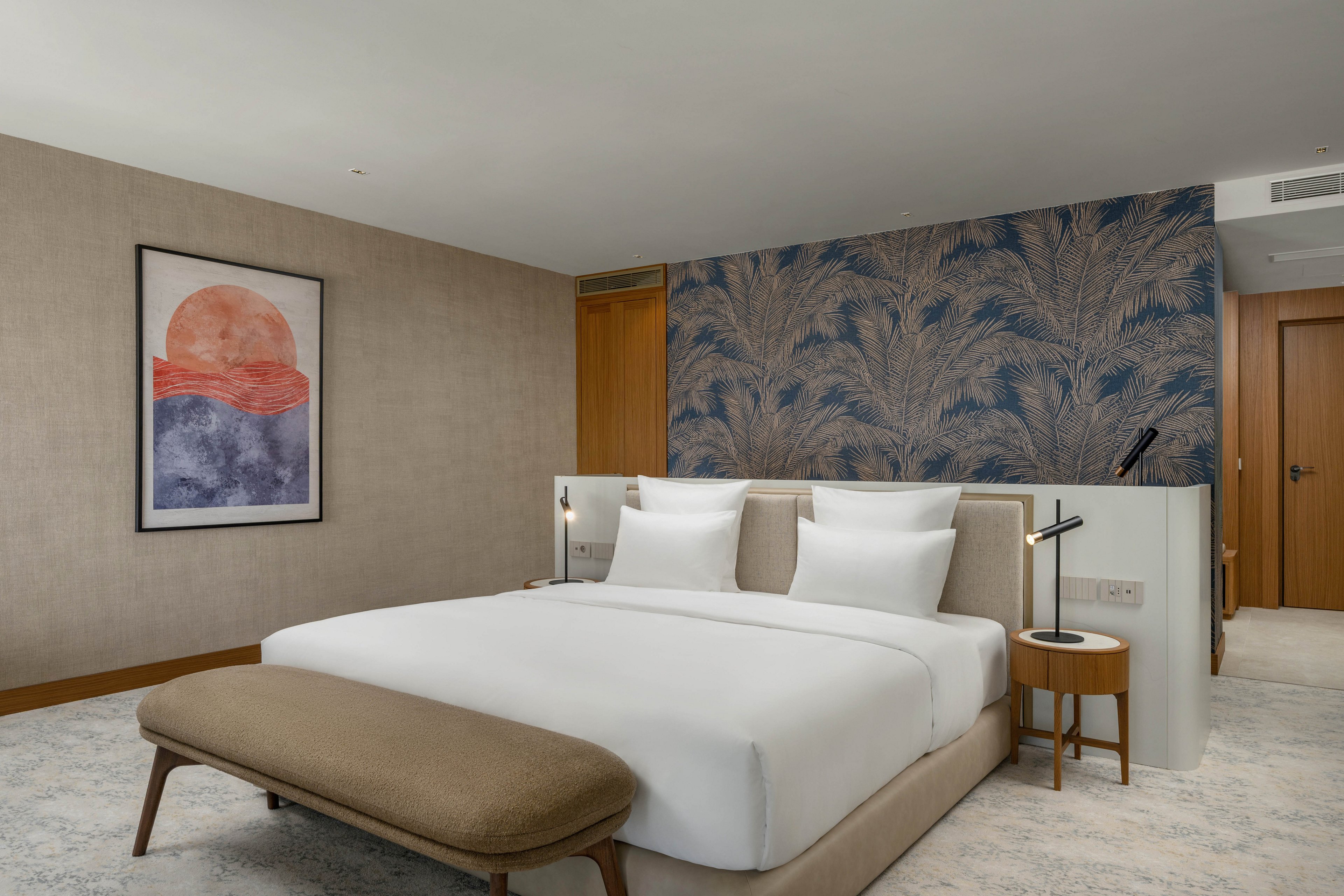 A contemporary hotel bedroom features a large bed with crisp white linens and a cushioned bench at the foot. The room includes a patterned accent wall, bedside tables with modern lamps, and neutral-toned carpeting. A framed abstract artwork with a sun and waves motif hangs on the opposite wall. Soft lighting and clean lines create a calm, minimalist atmosphere suitable for relaxation or business travel.
