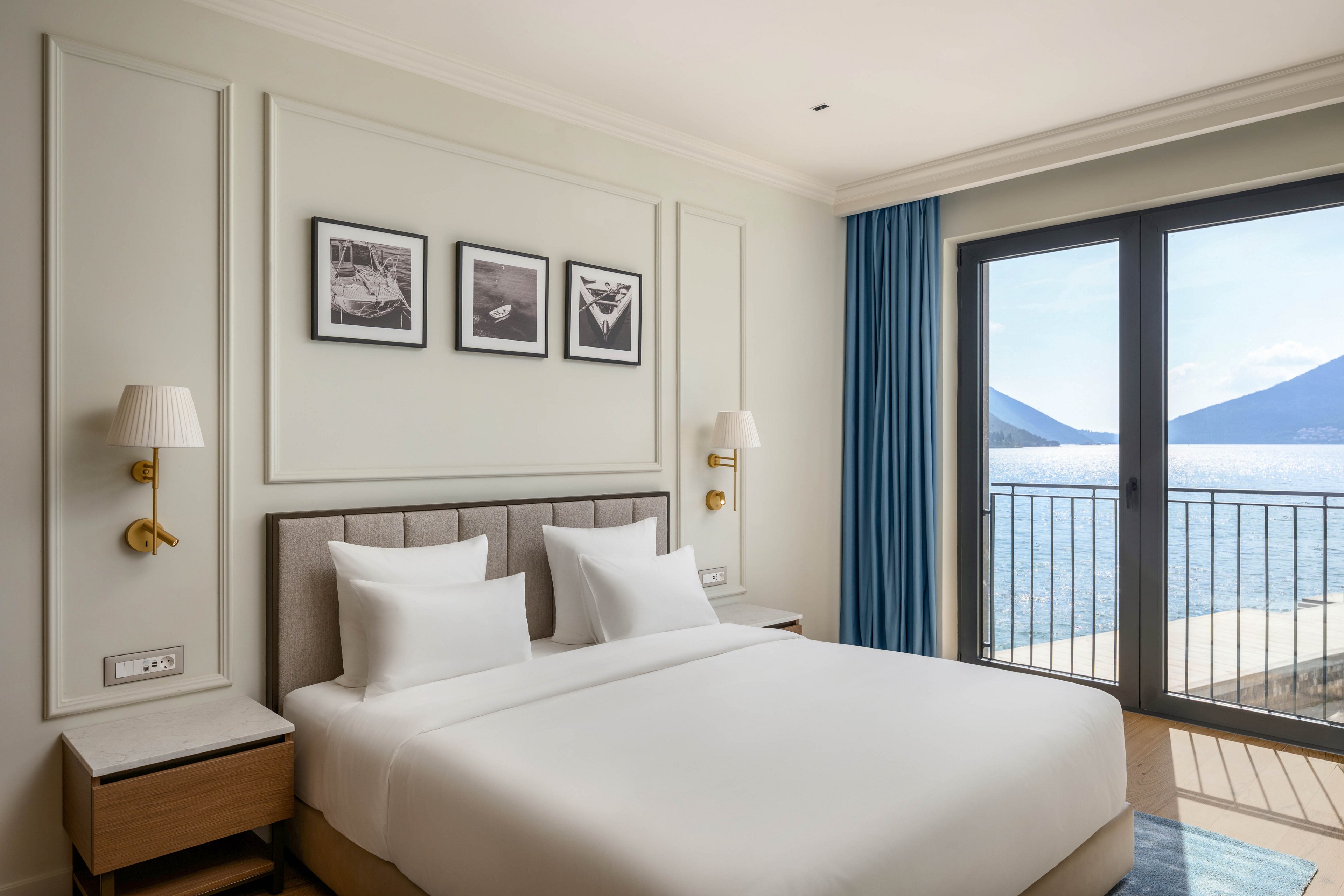 A neatly made double bed with crisp white linens is centered in a modern hotel bedroom. The room features light-colored walls, wall-mounted lamps, and framed black-and-white artwork above the headboard. Large glass doors open onto a balcony overlooking a calm ocean and distant mountains. Blue curtains frame the view, adding a subtle contrast to the neutral interior tones.