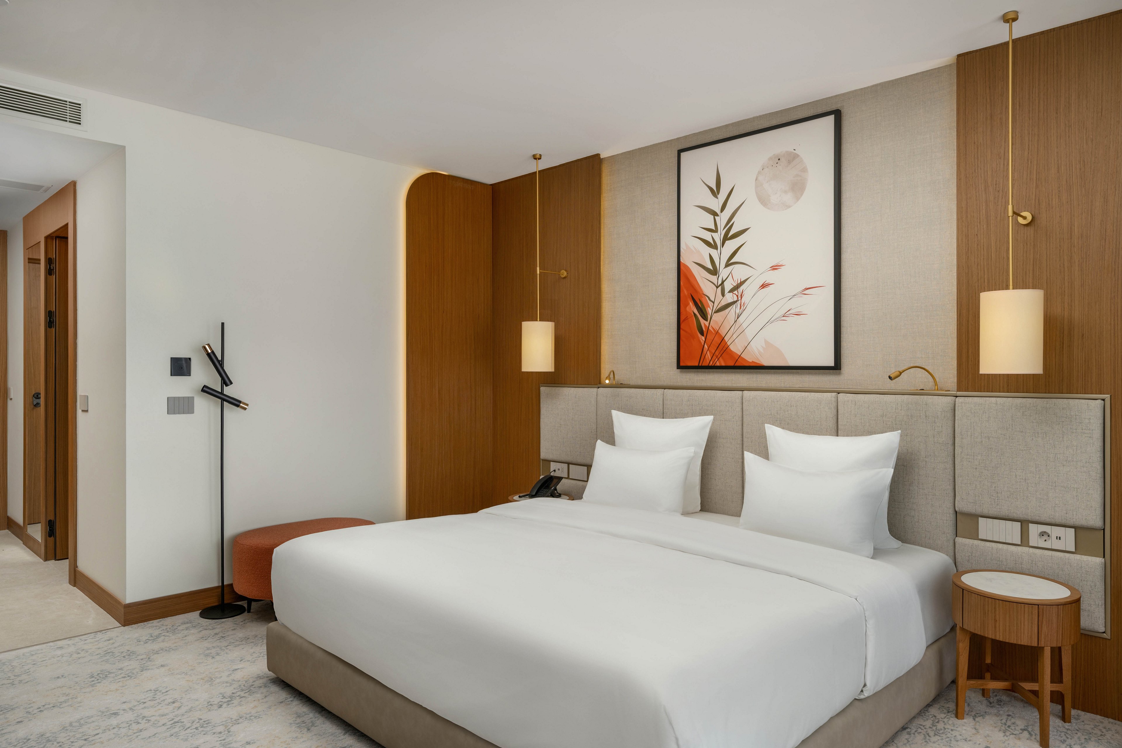 A contemporary hotel bedroom features a large neatly made bed with crisp white linens and padded headboard. Warm wooden panels, pendant lights, and a framed abstract artwork create a sophisticated focal point above the bed. A small round side table, upholstered stool, and sleek wall-mounted reading light add functional comfort. The neutral color palette and soft lighting give the room a calm, inviting atmosphere.