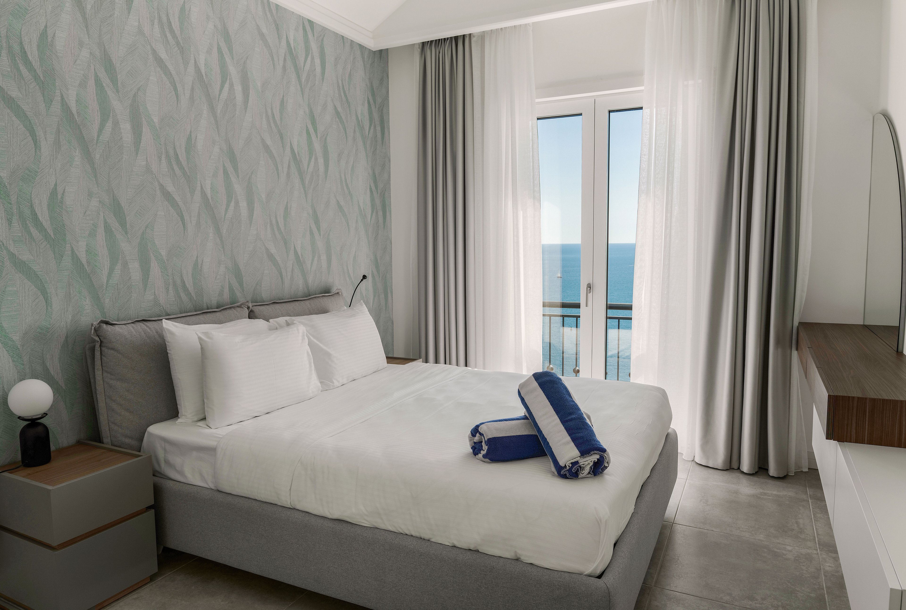 A contemporary bedroom features a neatly made double bed with white linens and blue accent towels placed on top. Large glass doors open to a balcony overlooking a calm ocean, framed by sheer white and gray curtains. The room includes minimalist bedside tables, a small spherical lamp, and a wall with subtle patterned wallpaper. Soft natural light enhances the clean, tranquil atmosphere and modern decor.