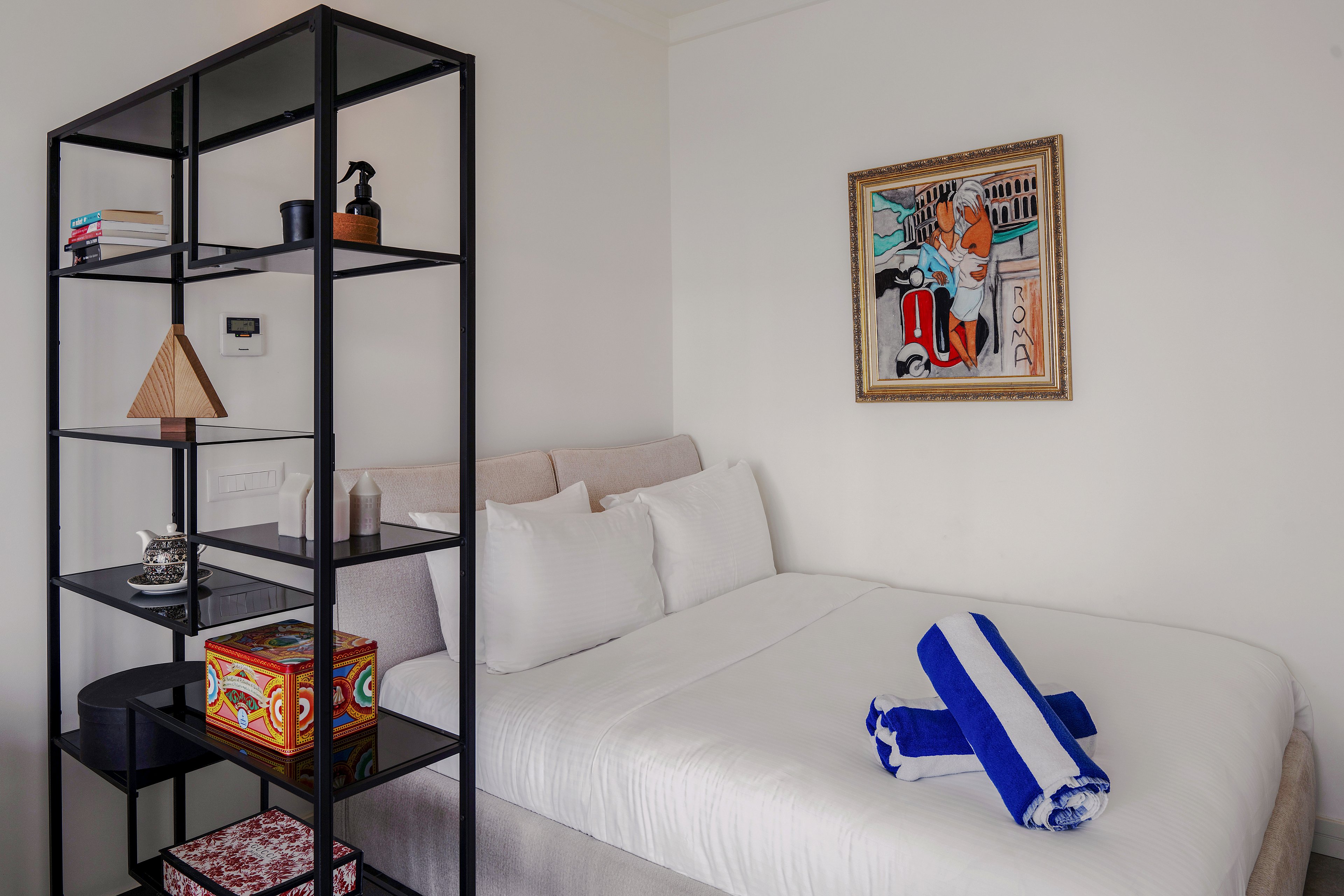 A neatly made bed with white linens sits beside a tall black metal shelving unit in a modern bedroom. Two rolled blue and white striped towels rest on the bed, suggesting a guest-ready space. The shelves display books, small sculptures, and colorful decorative boxes that add visual interest. A framed abstract painting hangs on the white wall above the bed, enhancing the contemporary and artistic atmosphere.