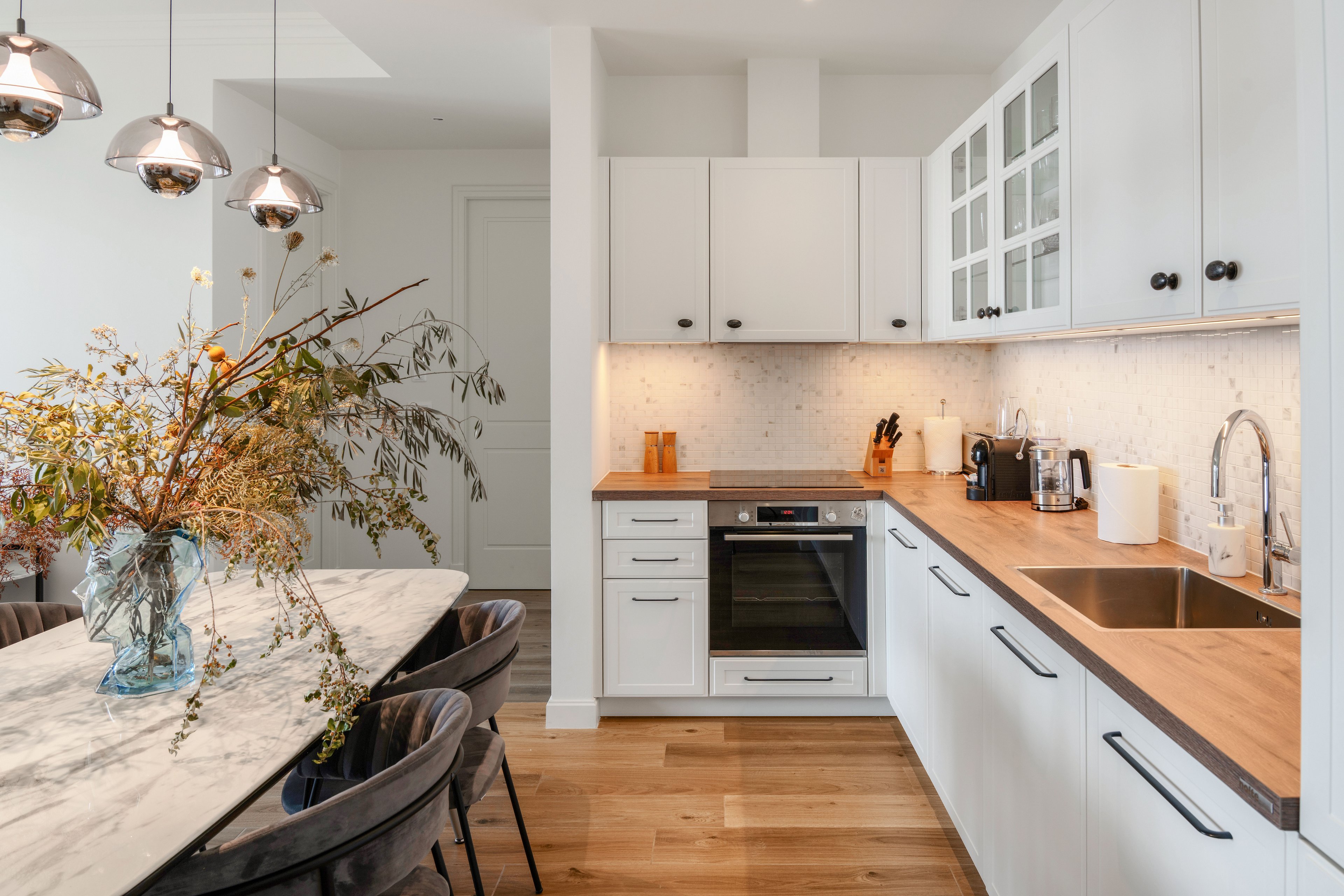 Bright contemporary kitchen interior featuring white cabinetry and warm wooden countertops. An integrated oven and stainless steel sink are built into a long counter with small appliances and utensils neatly arranged. Pendant lights hang above a marble dining table decorated with a large glass vase of dried flowers. Light wood flooring and soft under-cabinet lighting create a cozy, minimalist atmosphere.