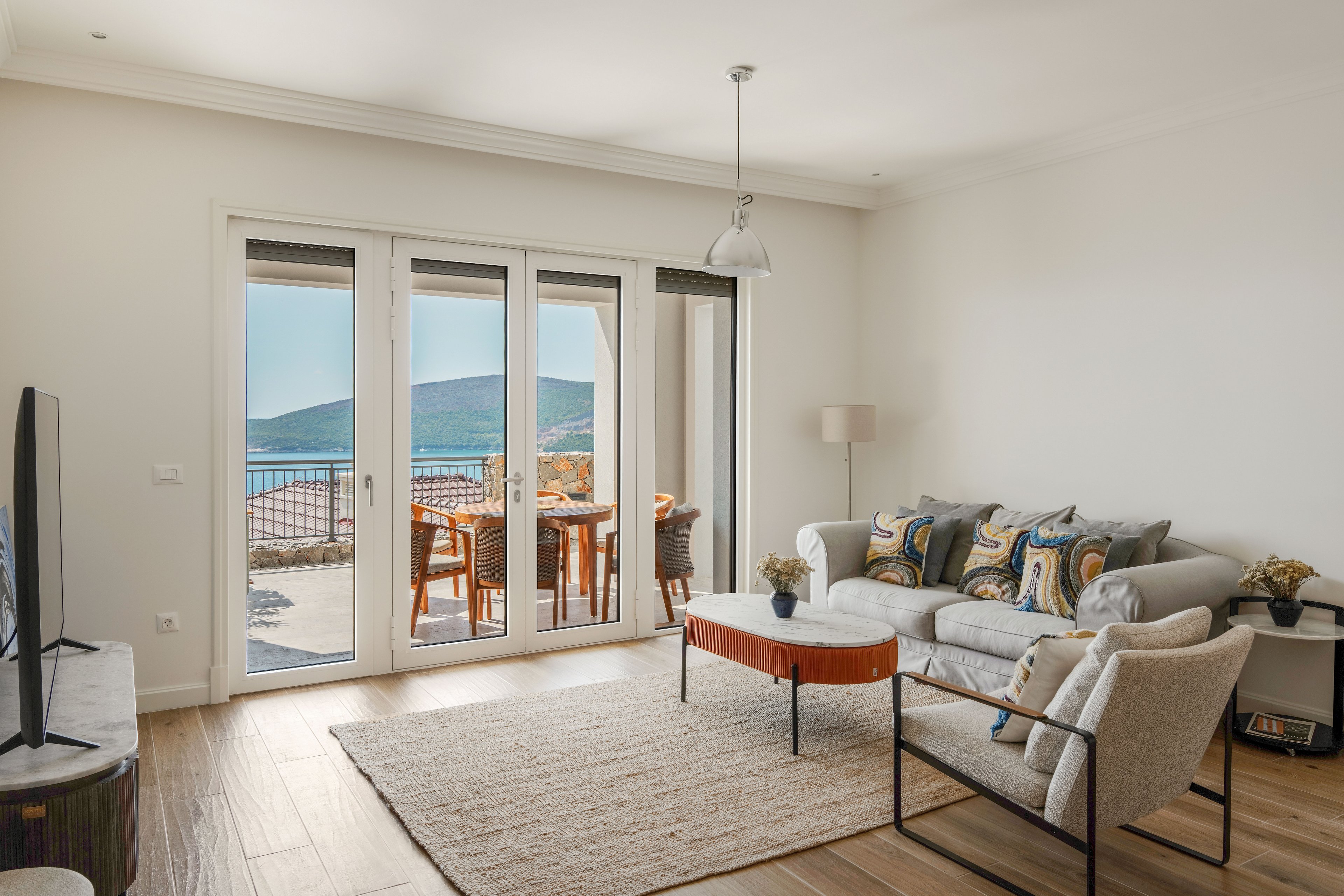 A modern living room features a light-colored sofa with patterned cushions and a simple coffee table on a neutral rug. Large glass doors open to a balcony with wooden outdoor furniture overlooking a scenic coastal landscape and distant hills. Warm wood flooring and soft beige tones create a relaxed, airy atmosphere. The space is neatly arranged, emphasizing comfort, natural light, and the ocean view.