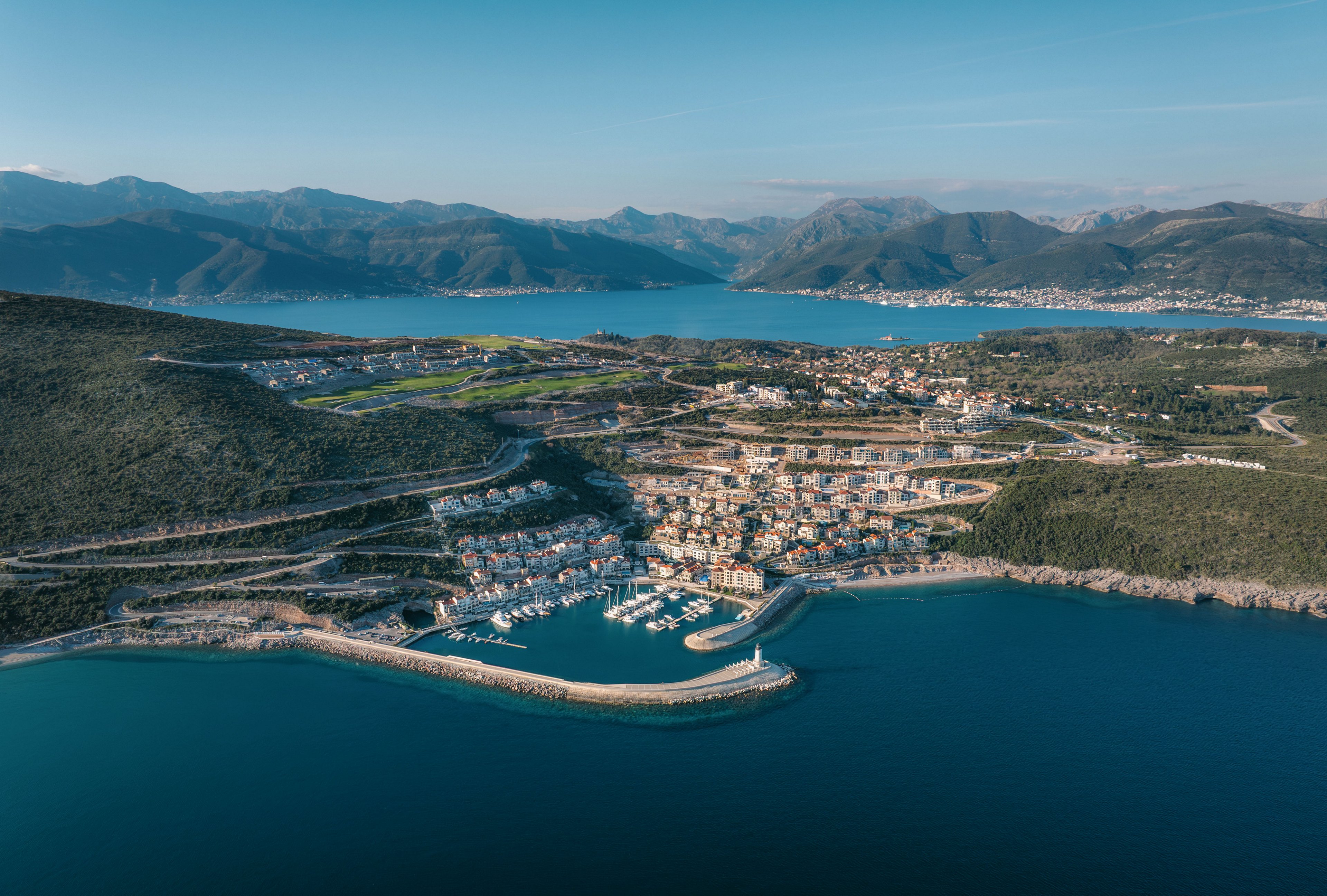 Aerial view of a coastal town with a marina extending into calm blue water. Residential buildings and streets spread across the hillside, bordered by green terrain and winding roads. The harbor features moored boats and a protective breakwater, with a lighthouse at the entrance. Distant mountains and additional bays frame the background under a clear sky.
