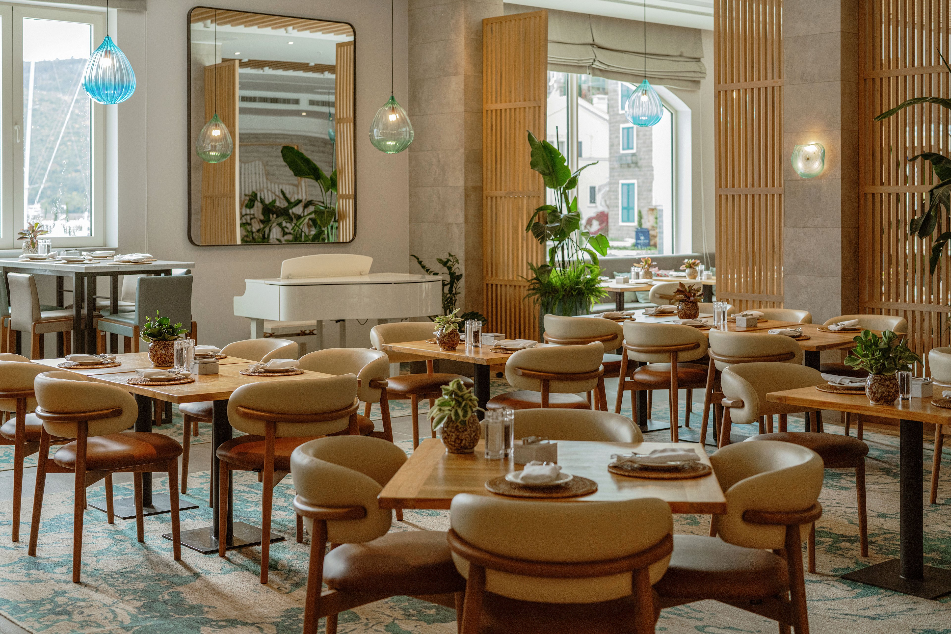 A modern restaurant dining area features neatly arranged wooden tables and cushioned chairs in neutral tones. The space is bright and airy, with large windows and decorative wooden room dividers. A white piano stands near the back wall, adding an elegant focal point beneath a large rectangular mirror. Pendant glass lights and small potted plants on each table contribute to a cozy, stylish atmosphere.