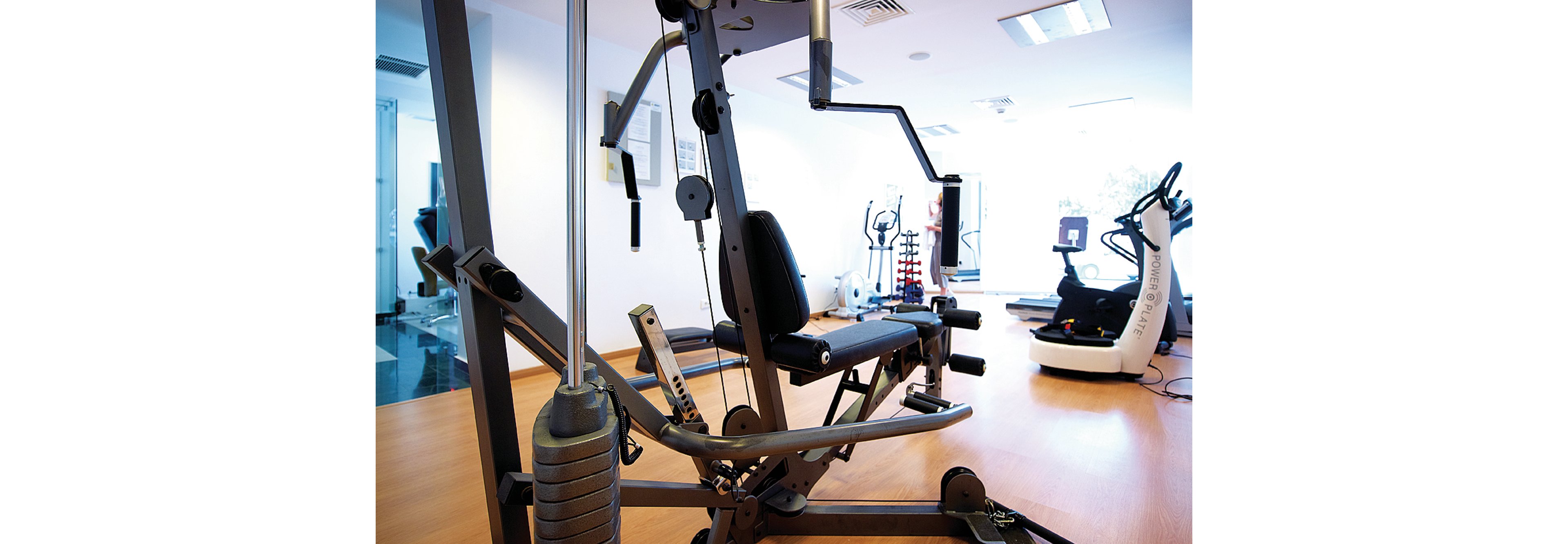 A multi-functional gym machine is prominently displayed in a well-lit fitness room. The setting includes additional exercise equipment such as stationary bikes and weights, creating a professional workout environment. The room features large windows allowing natural light to enhance the space. The equipment is designed for strength training and overall fitness routines.