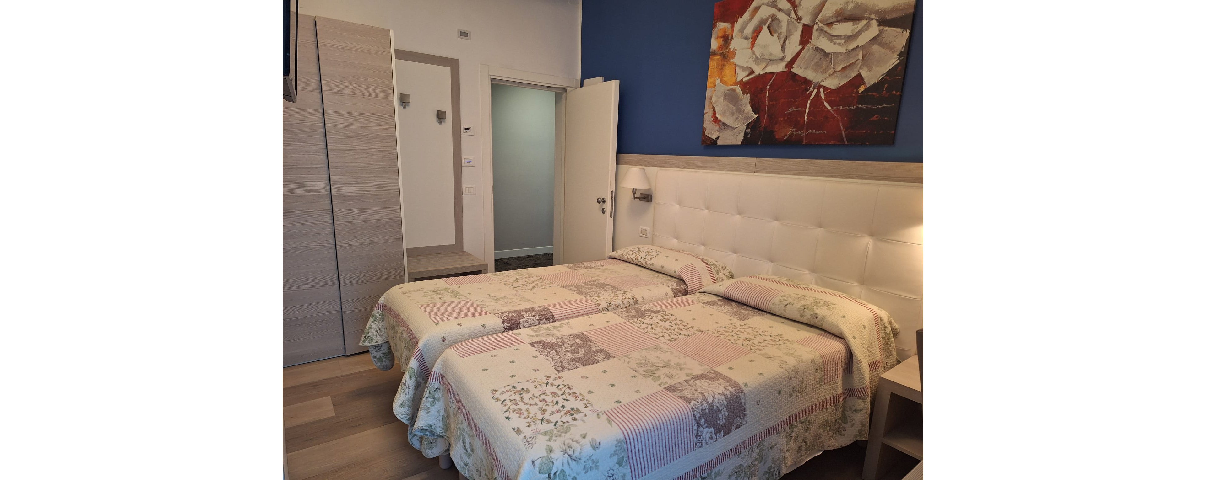 A neatly arranged bedroom features two twin beds with matching patchwork quilts in soft pastel colors. The room has a padded white headboard running along the wall and a bold blue accent wall above it. A large abstract painting hangs over the beds, adding a touch of contemporary art. Light wood flooring, a wardrobe, and an open door leading to a hallway complete the clean, modern interior.