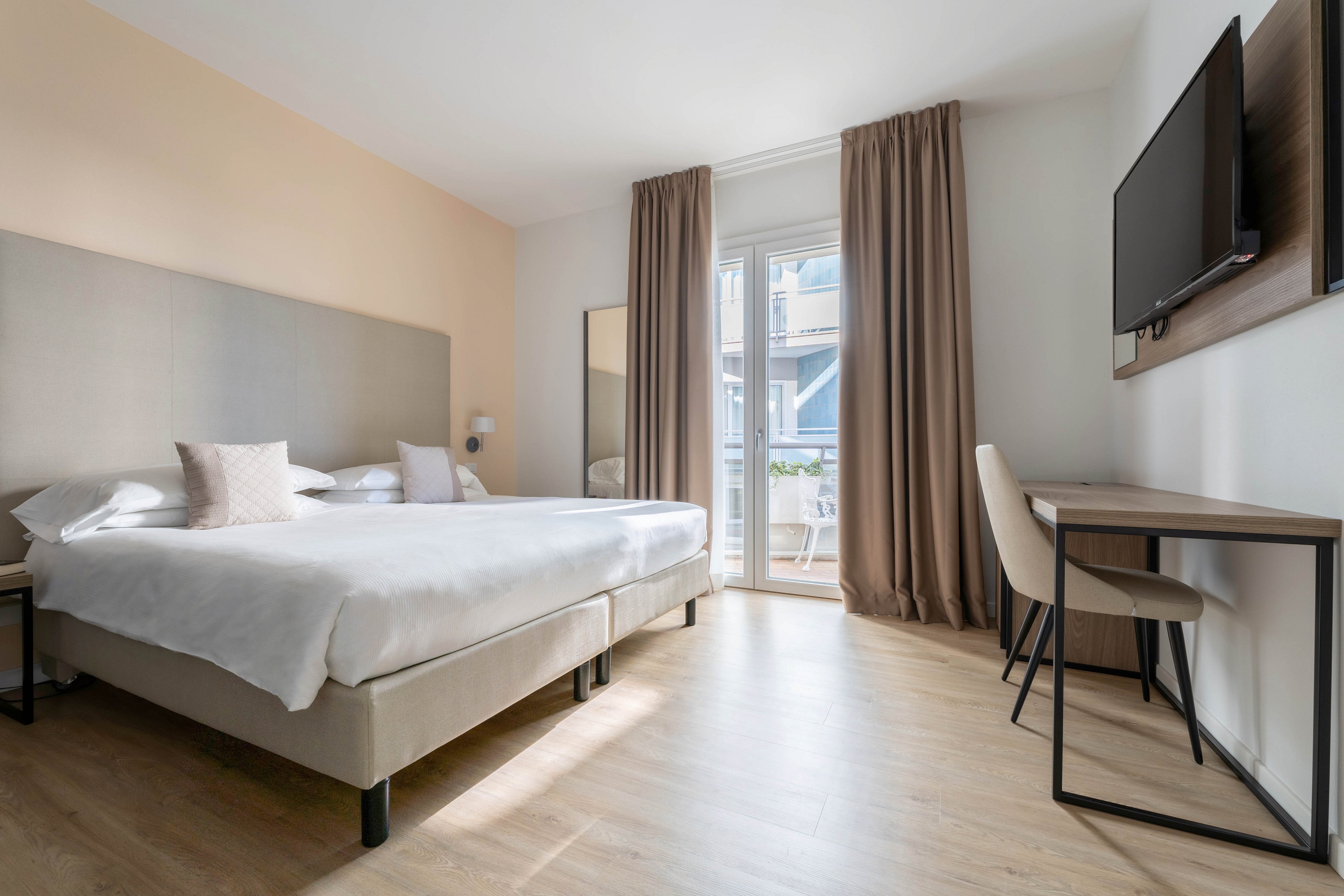 A bright, modern hotel bedroom features a large neatly made bed with white linens and neutral-toned pillows. Floor-to-ceiling curtains frame a glass door that opens onto a small balcony with outdoor seating. A wall-mounted flat-screen television faces the bed, and a simple desk with a chair sits along the side wall. Light wood flooring and soft beige accents create a clean, minimalist atmosphere.