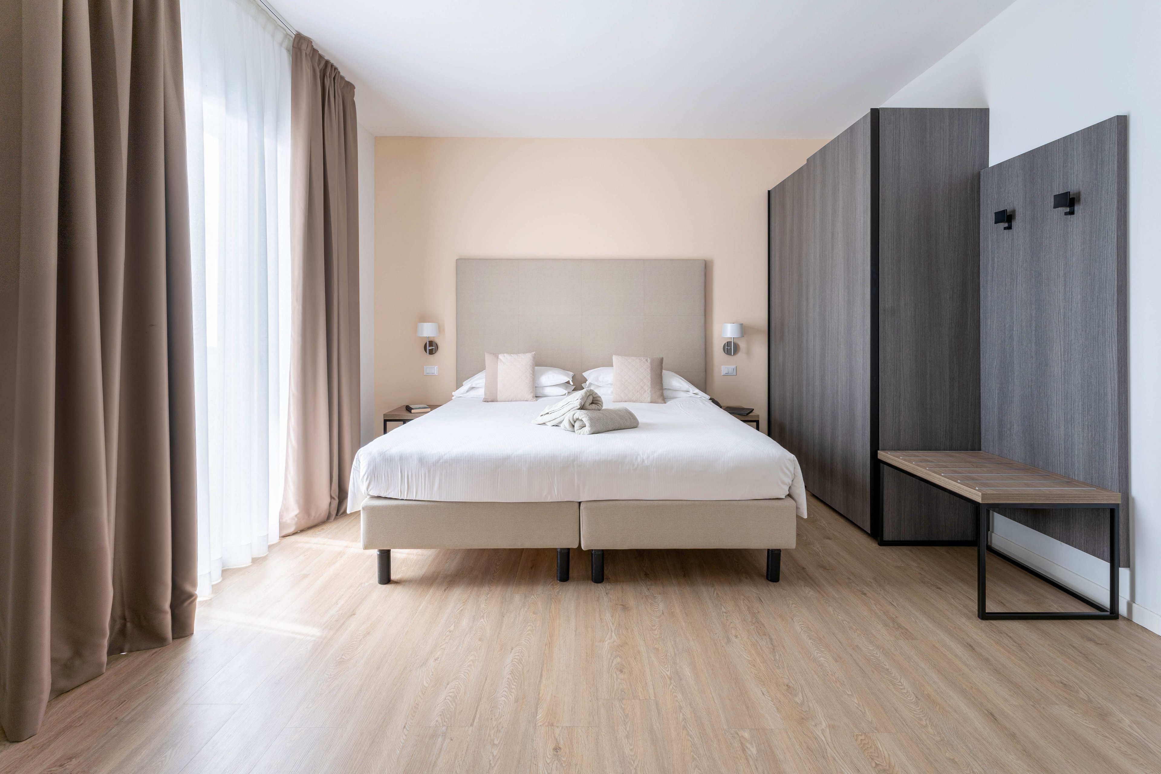 A modern hotel bedroom features a large double bed with neutral bedding and neatly arranged pillows. The room has light wood flooring, a soft beige accent wall, and floor-to-ceiling curtains framing a bright window. Dark wardrobes and a simple bench add contrast to the minimalist decor. Overall, the space feels clean, spacious, and inviting with a calm, contemporary style.