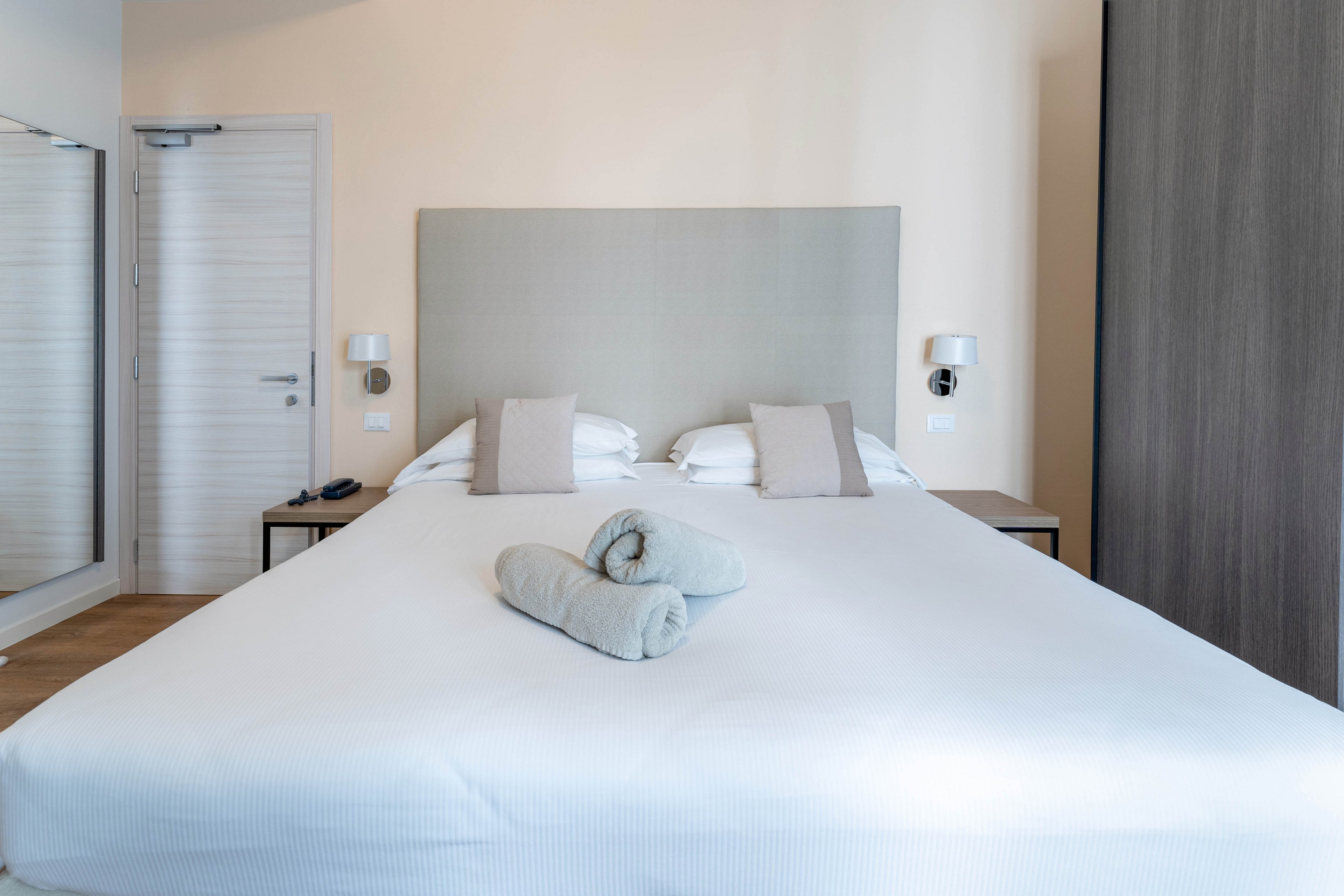 A contemporary hotel bedroom features a large neatly made bed with a light upholstered headboard. Two rolled towels are placed at the center of the bed, adding a welcoming touch. Matching bedside tables with wall-mounted lamps flank the bed, and a wardrobe and mirror are visible in the room. The neutral color palette and clean lines create a calm, minimalist atmosphere.