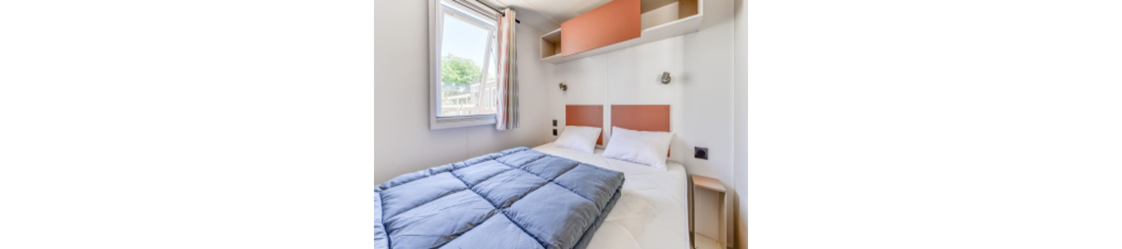 A neatly arranged bedroom features a double bed with white pillows and a blue comforter. The room is well-lit by natural light coming through a window with pastel curtains, and there are built-in shelves above the bed. The walls are white, creating a clean and airy atmosphere, while the decor is minimal and modern.