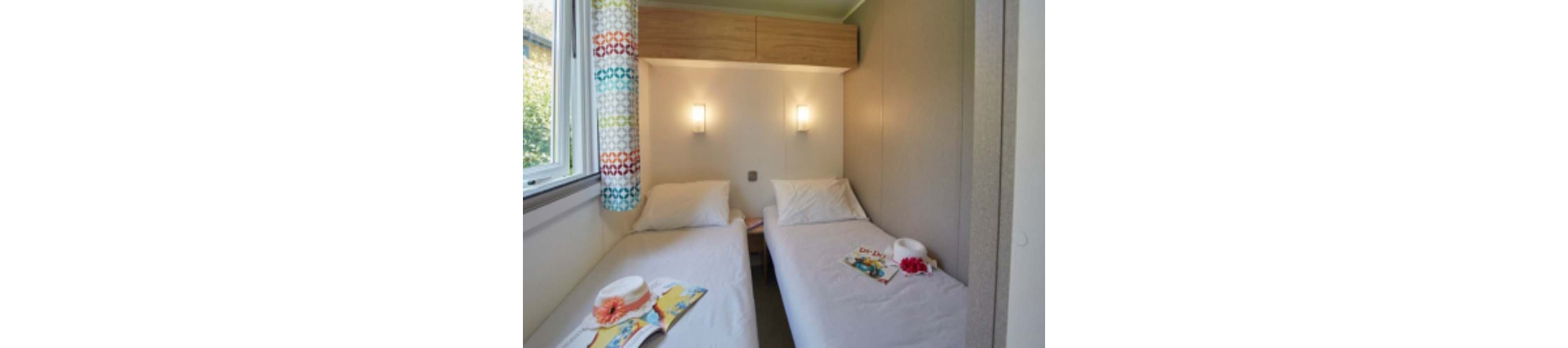 This image shows a compact bedroom featuring two single beds with white linens, separated by a small nightstand. The room is brightly lit by two wall-mounted lights and natural daylight from a window with multicolored patterned curtains. On each bed, there is a white hat and a magazine, adding a casual and inviting touch. The walls are neutral, and overhead storage cabinets provide additional functionality.