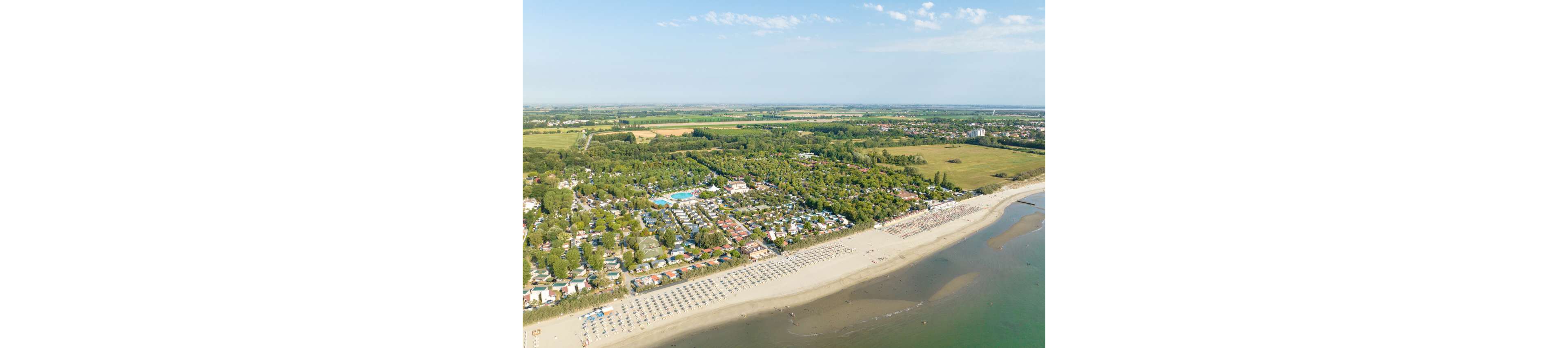 This aerial photograph captures a sprawling beachfront resort adjacent to a wide sandy beach. Rows of umbrellas and sun loungers line the shore, while lush greenery and pools are visible within the resort grounds. The setting is tranquil, with open fields and distant buildings in the background. The image conveys a relaxed, summery mood with clear skies and gentle waves. No visible text or numeric values are present in the image.