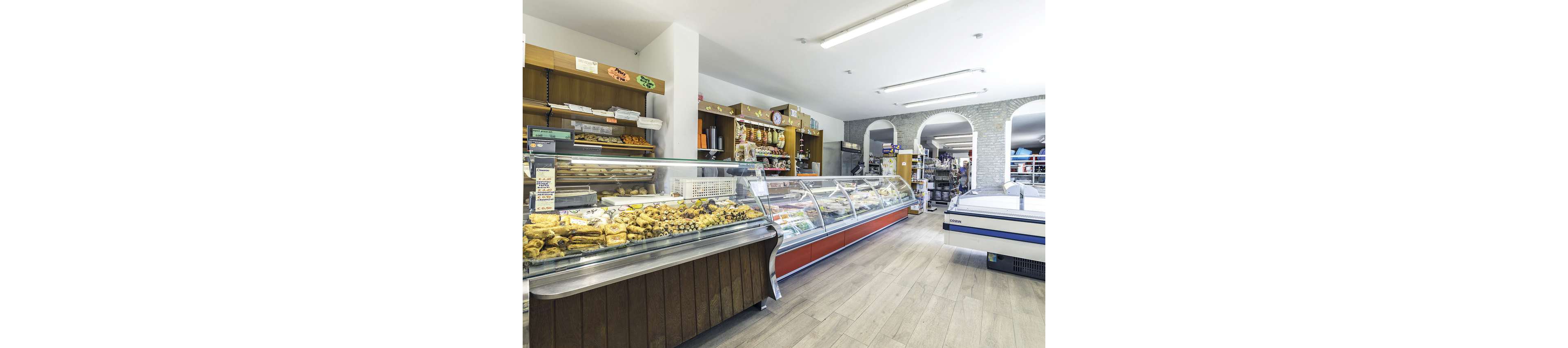 A well-lit delicatessen features a long refrigerated display case filled with assorted pastries, meats, and cheeses. Shelves behind the counter are stocked with packaged goods and bread, while the open layout reveals more products in the background. The space has light wooden flooring and white walls, creating a clean and inviting atmosphere. No visible people are present, and no explicit numbers or brand names are clearly identifiable.