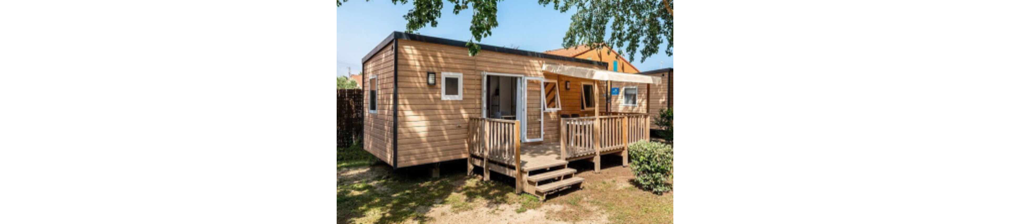A contemporary wooden mobile home is shown with a small front porch and steps leading up to the entrance. The setting is outdoors, surrounded by trees and greenery, with sunlight casting shadows on the ground. The home features large windows, a shaded awning, and a relaxed, inviting atmosphere. No visible text or numbers are present in the image.