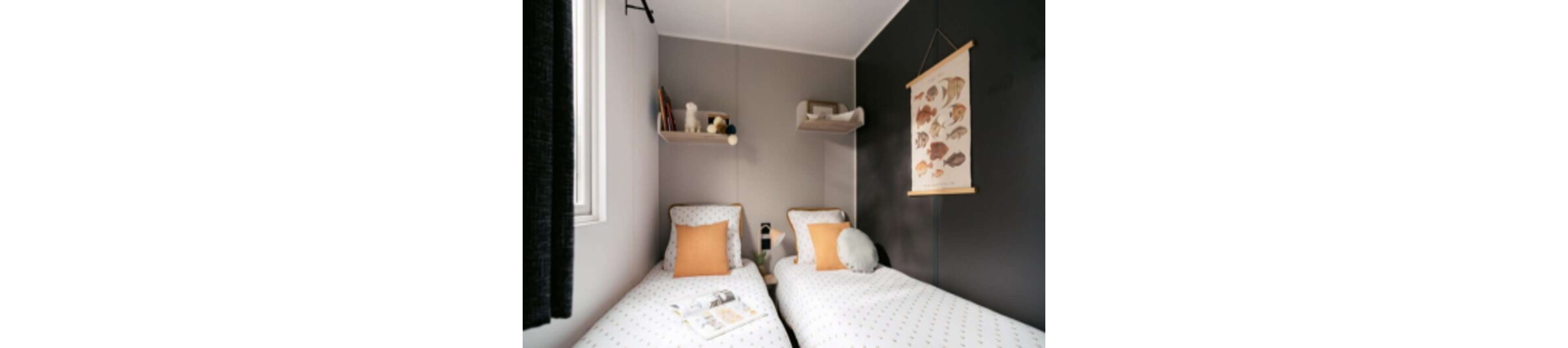 This image features a small bedroom with two twin beds, each adorned with white and dotted bedding and orange accent pillows. The room has a minimalist style, with shelves holding decorative items and a wall art print of various fish species. Soft natural light enters from a window, creating a calm and inviting atmosphere. No visible numbers or text are present in the image.