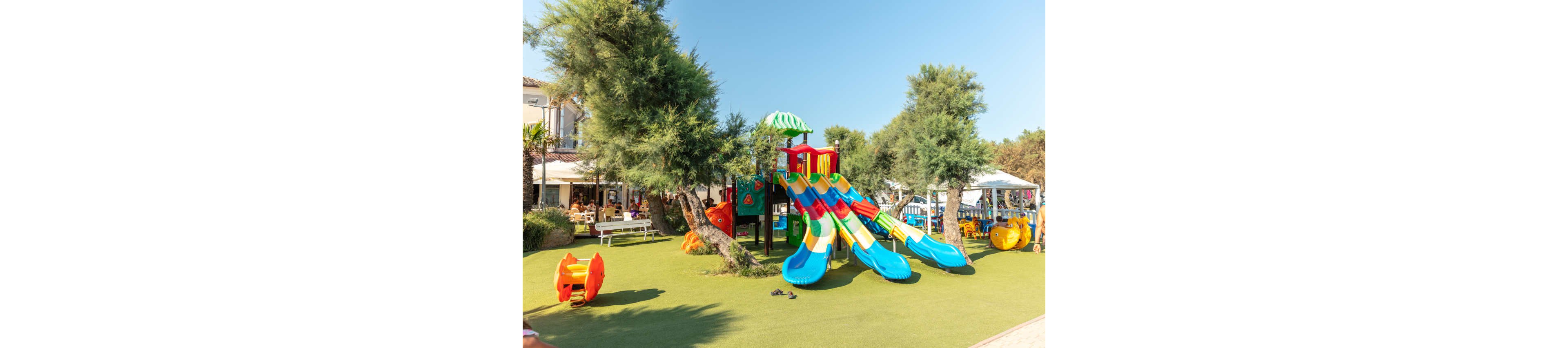 A vibrant children's playground features several bright blue and yellow slides under a green-striped canopy. The play structure is set on artificial grass, surrounded by trees and shaded seating areas. Families and children are visible in the background, enjoying a sunny day in a park-like setting. The overall mood is cheerful and lively, with clear skies and playful visuals.