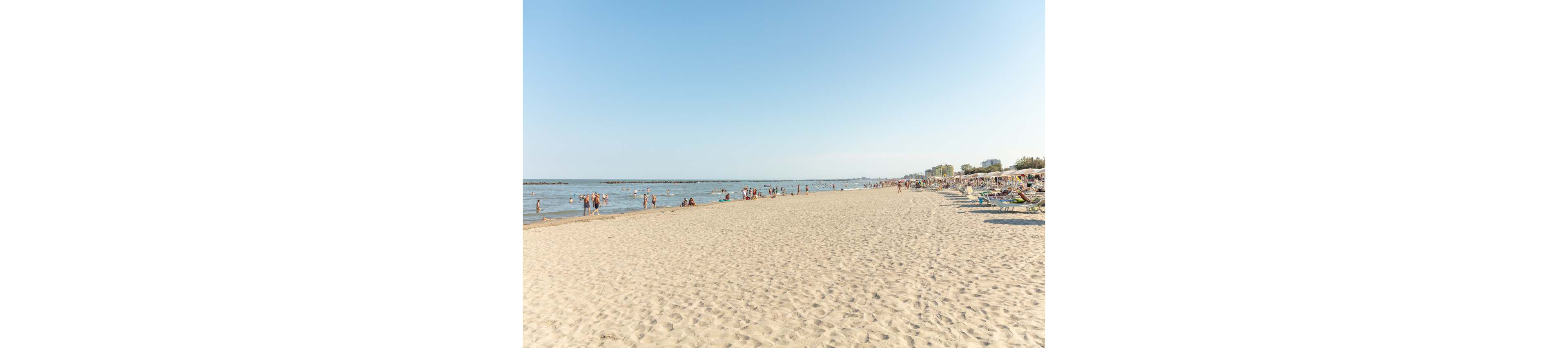 A wide sandy beach stretches along the coastline under a clear blue sky. Groups of people of various ages are seen relaxing on the sand and swimming in the calm sea. Rows of sun loungers and umbrellas line the right side, while distant buildings are visible in the background. The atmosphere is bright and leisurely, evoking a summer vacation mood. No visible text or numbers are present in the image.