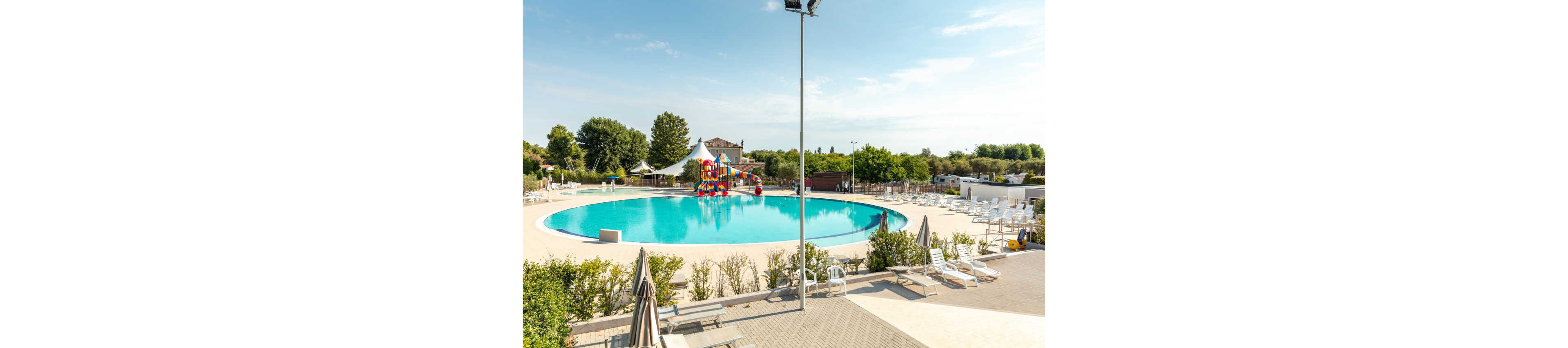 A large, round outdoor swimming pool is the main focus, surrounded by lounge chairs and umbrellas. In the background, a colorful water slide structure is visible, adding a playful element to the scene. The pool area is set in a landscaped environment with trees and greenery, under a bright blue sky. The overall mood is relaxed and inviting, ideal for leisure and recreation.