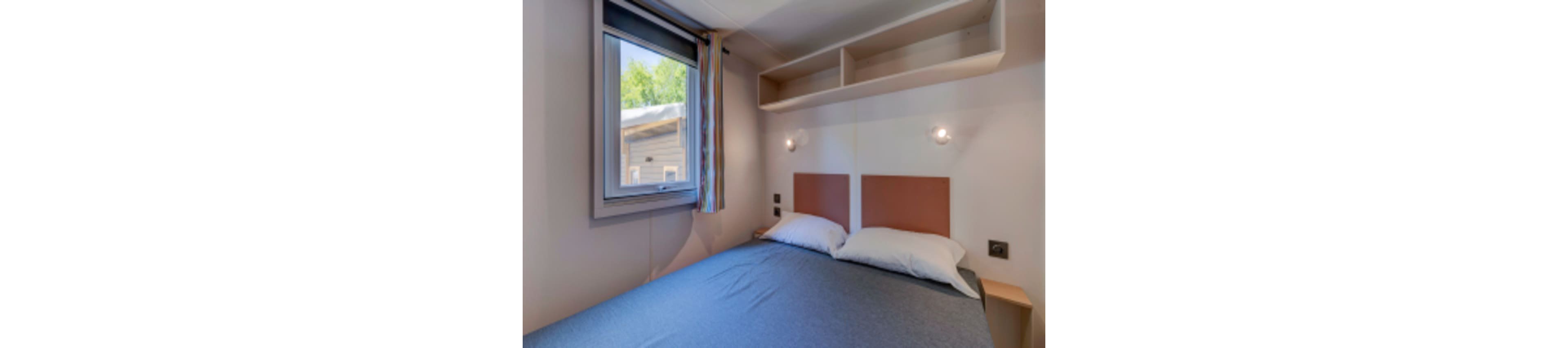 A compact bedroom features a double bed with two pillows and a blue-gray bedspread. The room has a window with colorful curtains, built-in shelving above the bed, and two wall-mounted reading lights. Natural light enters, highlighting the simple and functional decor. The setting suggests a peaceful, minimalist atmosphere.