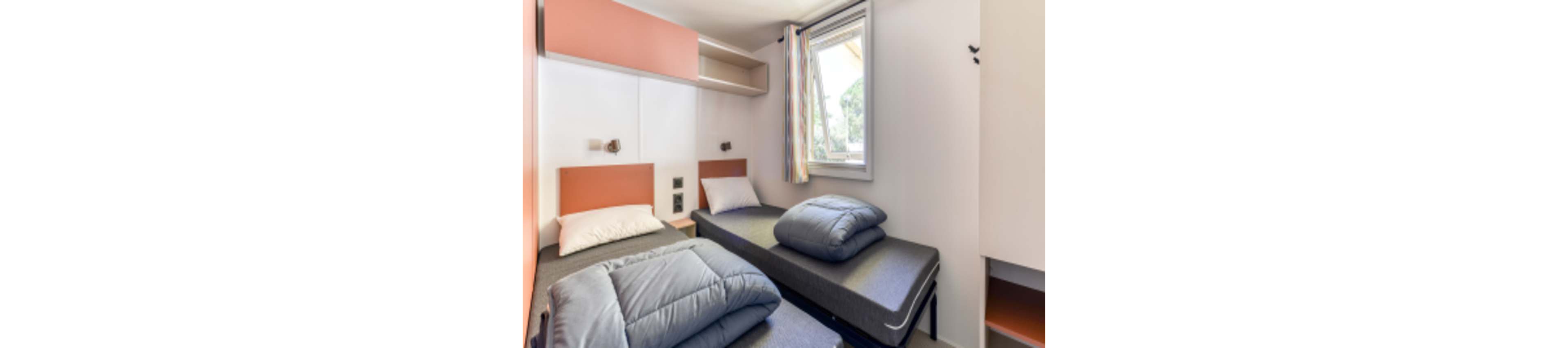 A compact twin bedroom features two single beds with gray bedding and orange headboards. Natural light enters through a window with colorful curtains, illuminating the minimalist interior. The room includes built-in shelving, a small closet, and neutral wall colors for a clean, modern look. No visible text or numbers are present in the image.