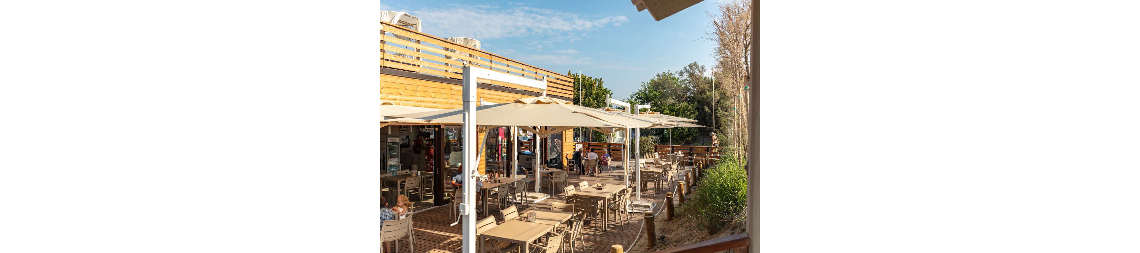 A spacious outdoor café features wooden deck seating under large cream umbrellas. Several people are seated at tables, enjoying a relaxed atmosphere in the sunshine. The setting includes natural greenery, wooden railings, and a clear blue sky, creating a casual and inviting mood. The café structure is modern with light wood paneling and open-air dining. No visible text or numbers are present in the image.