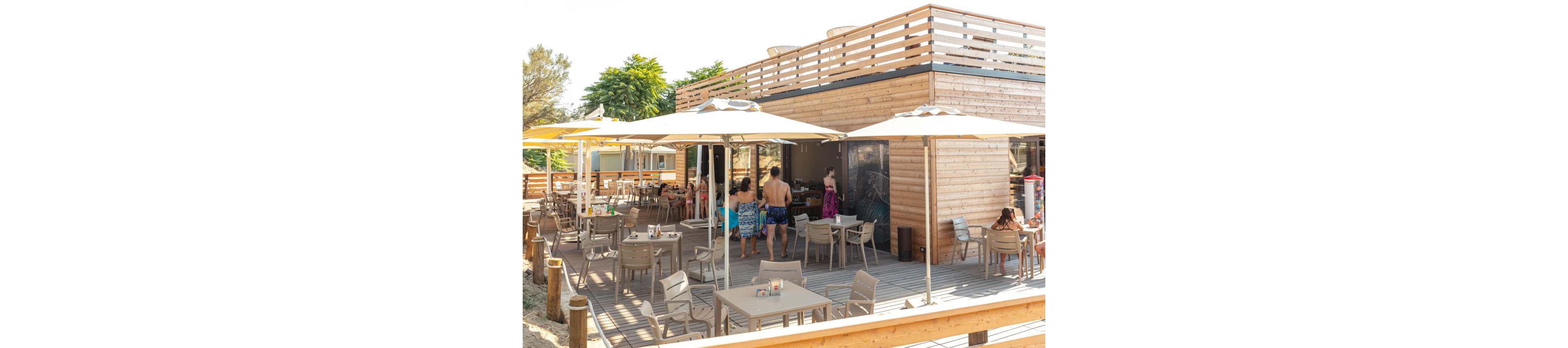 A group of people in swimwear gather at a modern wooden café with outdoor seating under large umbrellas. The setting is bright and sunny, with a relaxed summer atmosphere and light wood tones throughout. Several tables and chairs are arranged on the deck, and lush greenery surrounds the area. The scene captures a casual, social moment at a beachside eatery.
