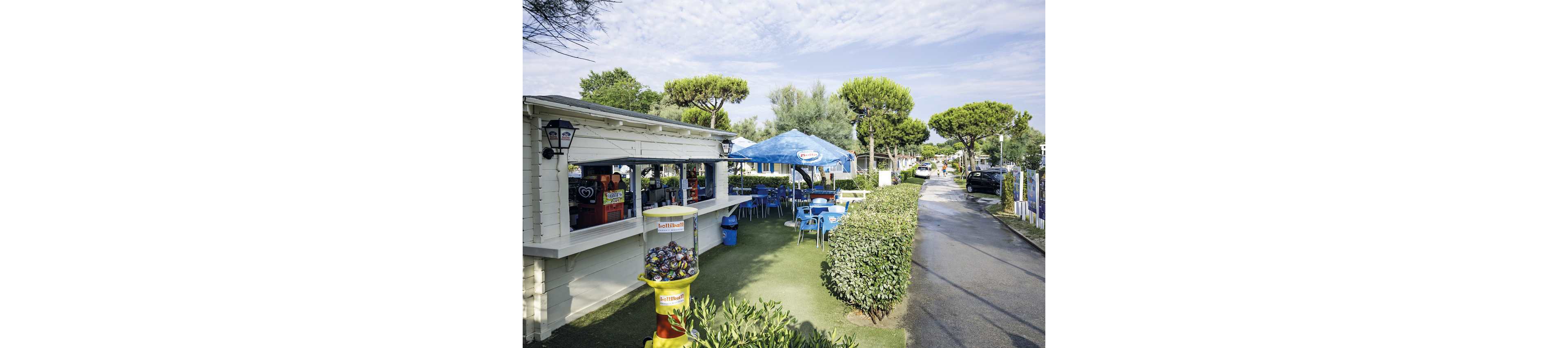 A small outdoor snack bar is set in a green, landscaped area with several blue tables and chairs under a large branded umbrella. A vending machine filled with colorful balls stands in the foreground, displaying the text 'Autoball.' The setting is bright and casual, with a relaxed summer atmosphere and a few people visible behind the counter.