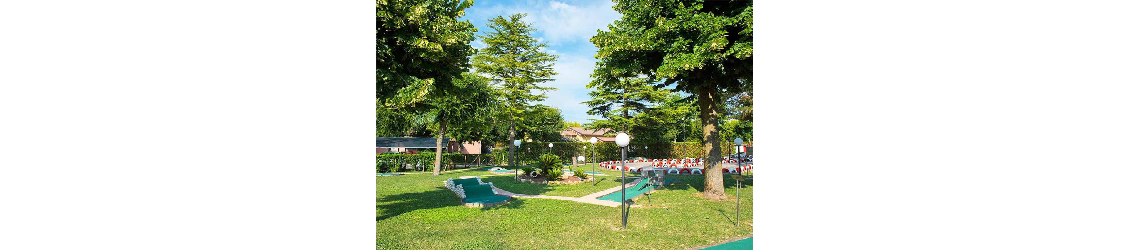 A vibrant outdoor mini golf course is set amidst well-maintained grass and tall leafy trees. The scene features several mini golf holes with green turf, bordered by white stones and surrounded by decorative lampposts. In the background, there are red and white seating areas and residential buildings, all under a bright blue sky with scattered clouds.
