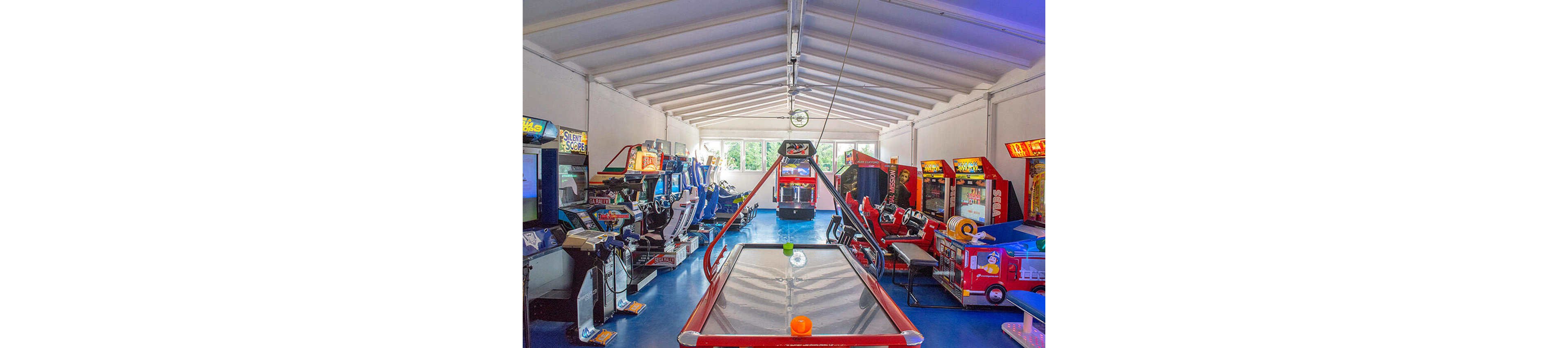 A spacious arcade room features a central air hockey table with orange and green pucks. The setting is filled with various classic arcade game machines, including racing and claw games, arranged along the walls. The room is brightly lit with natural light streaming through large windows, creating a lively and playful atmosphere. The blue floor and white ceiling enhance the vibrant, energetic mood.
