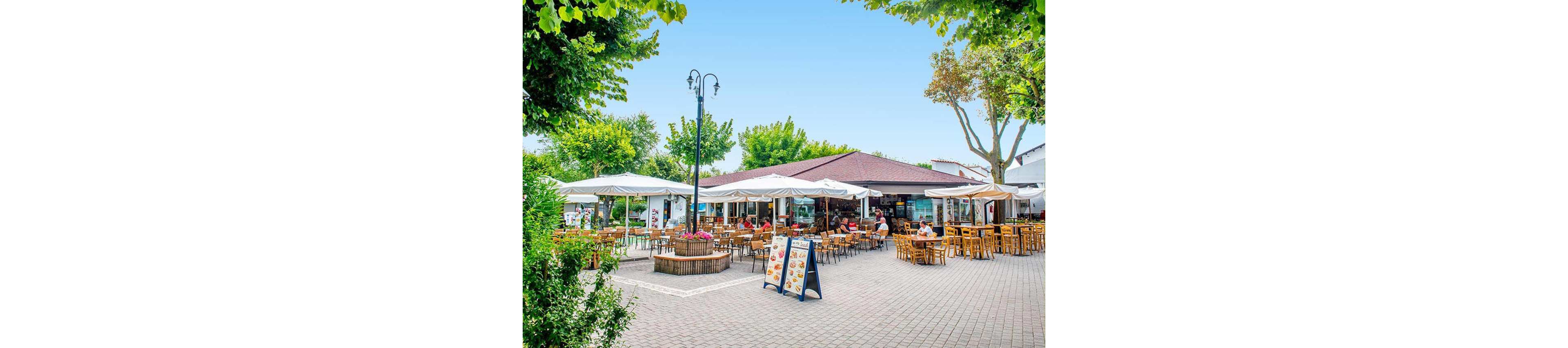An open-air café features numerous wooden tables and chairs arranged under large white umbrellas. The setting is a sunny, tree-lined plaza with a relaxed atmosphere, and a prominent menu board stands at the entrance. Several people are seated, enjoying food and drinks in a casual, social environment. The scene is bright with clear blue skies and lush greenery surrounding the area.