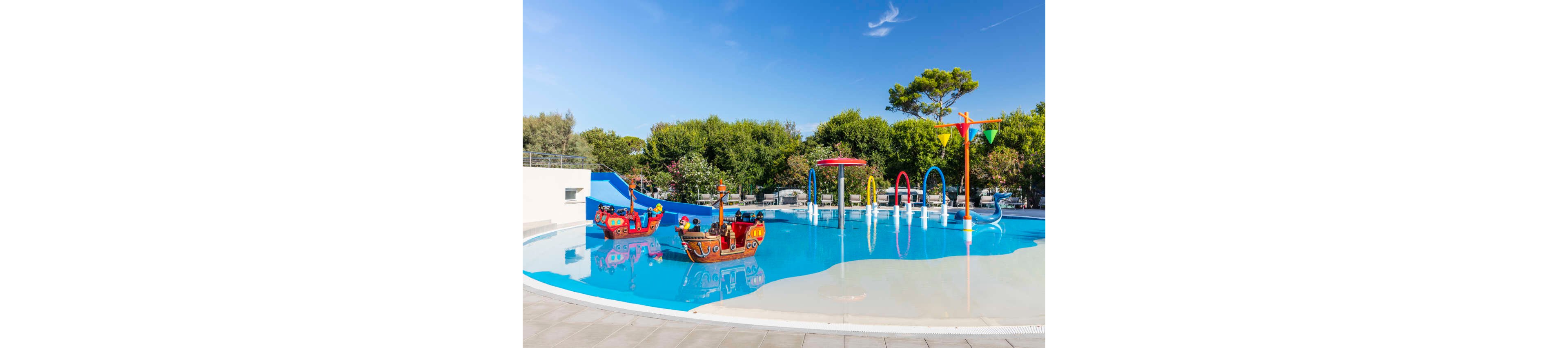 The main subject is a vibrant children's splash pool featuring pirate ship play structures and interactive water elements. The setting is outdoors, surrounded by lush greenery and clear blue skies, creating a playful and inviting atmosphere. Key visuals include colorful slides, water arches, and decorative flags, all designed for young children. No visible text or numeric values are present in the image.
